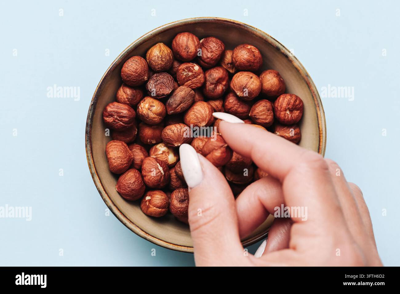 Hand wählt ganze Haselnüsse aus einer runden Schüssel auf hellblauem Hintergrund. Natürliche Lebensmittelkomposition mit Herbstnüssen und nahrhaften Snacks. Stockfoto
