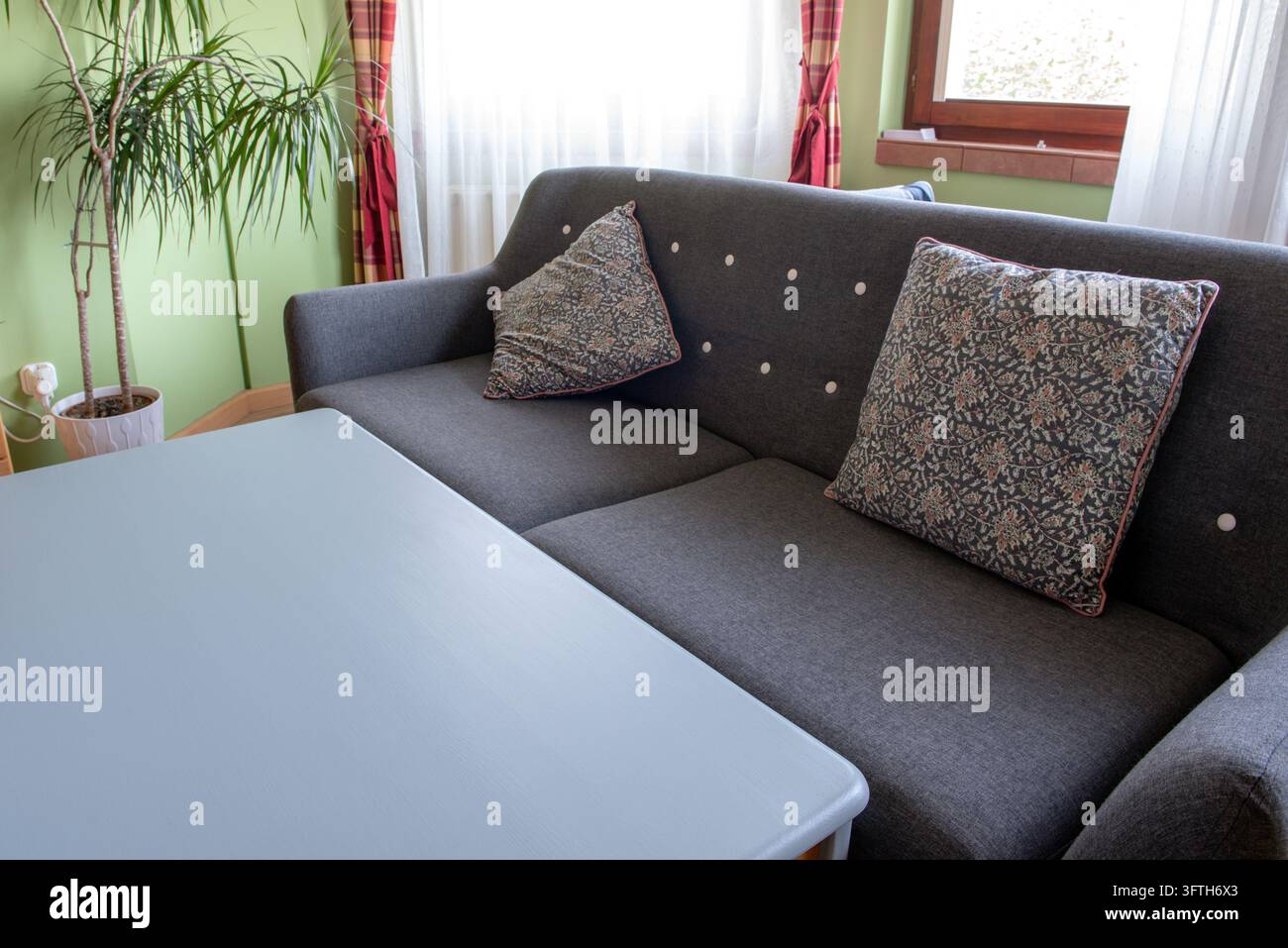 Ein graues gepolstertes Sofa mit floral gemusterten Kissen steht neben einem blauen Tisch in einem gemütlichen Wohnzimmer, mit einer Topfpflanze, durchsichtigen Vorhängen und einem Stockfoto
