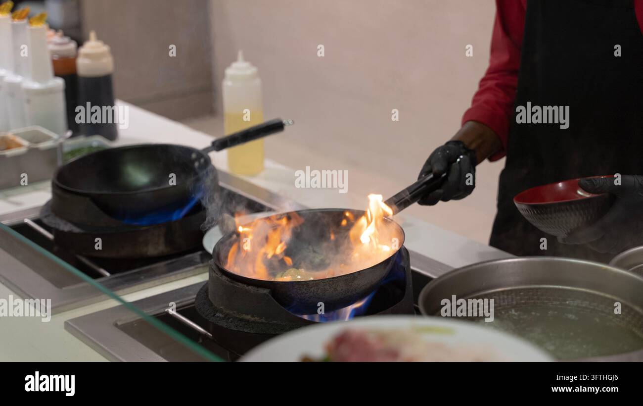 Professioneller Koch bereitet Essen in einer Bratpfanne über einer offenen Gasflamme in einer Großküche zu, wobei er Metallzangen verwendet und eine Servierschüssel hält. Stockfoto