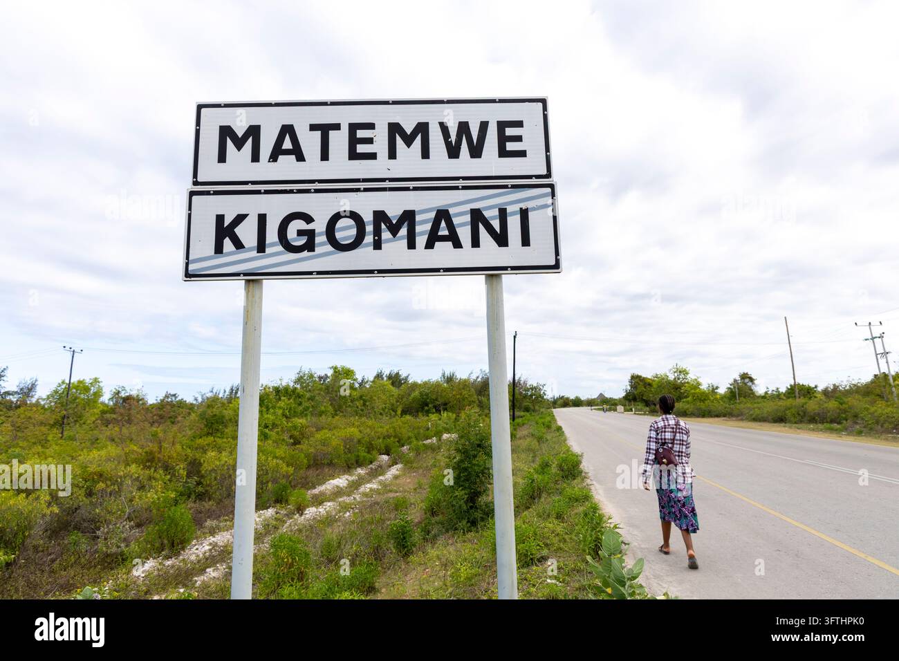 Frau mit geflochtenem Haar und einer kleinen Tasche geht an einer Straße vorbei an einem Dorfgrenzschild von Matemwe Kigomani unter einem bewölkten Himmel, Matemwe, Tansania Stockfoto