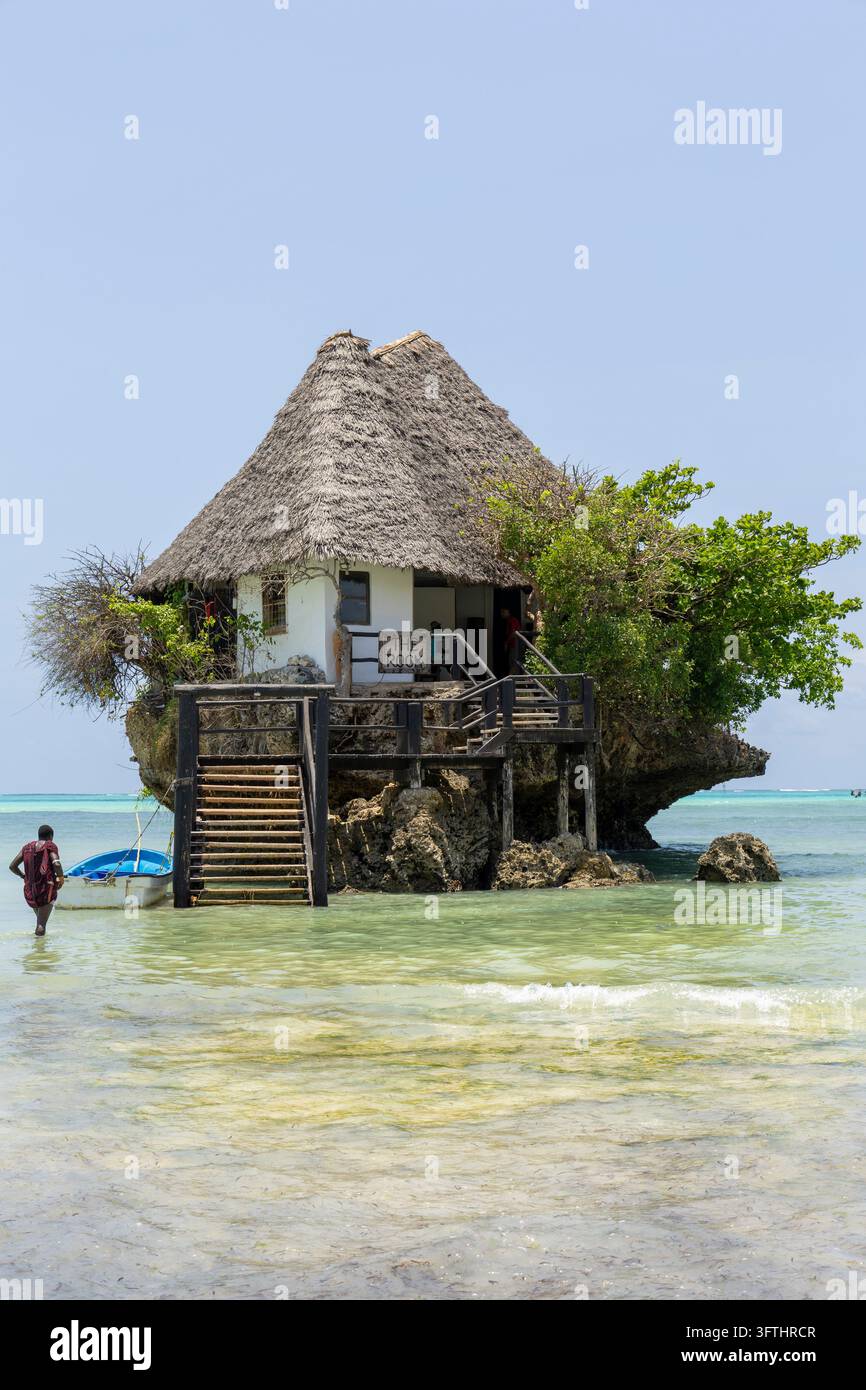 Strohgedecktes Restaurant The Rock auf einem Korallenfelsen im türkisblauen Indischen Ozean mit einer Holztreppe und einem kleinen Boot, Jambiani, Tansania Stockfoto