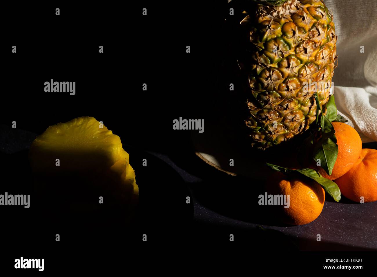Ananas auf einem rustikalen Holzblock mit Mandarinenscheiben, die darum gestapelt sind, neben geschnittenem Ananas auf einem schwarzen Teller vor dunklem Hintergrund. Dunkle Früchte Stockfoto