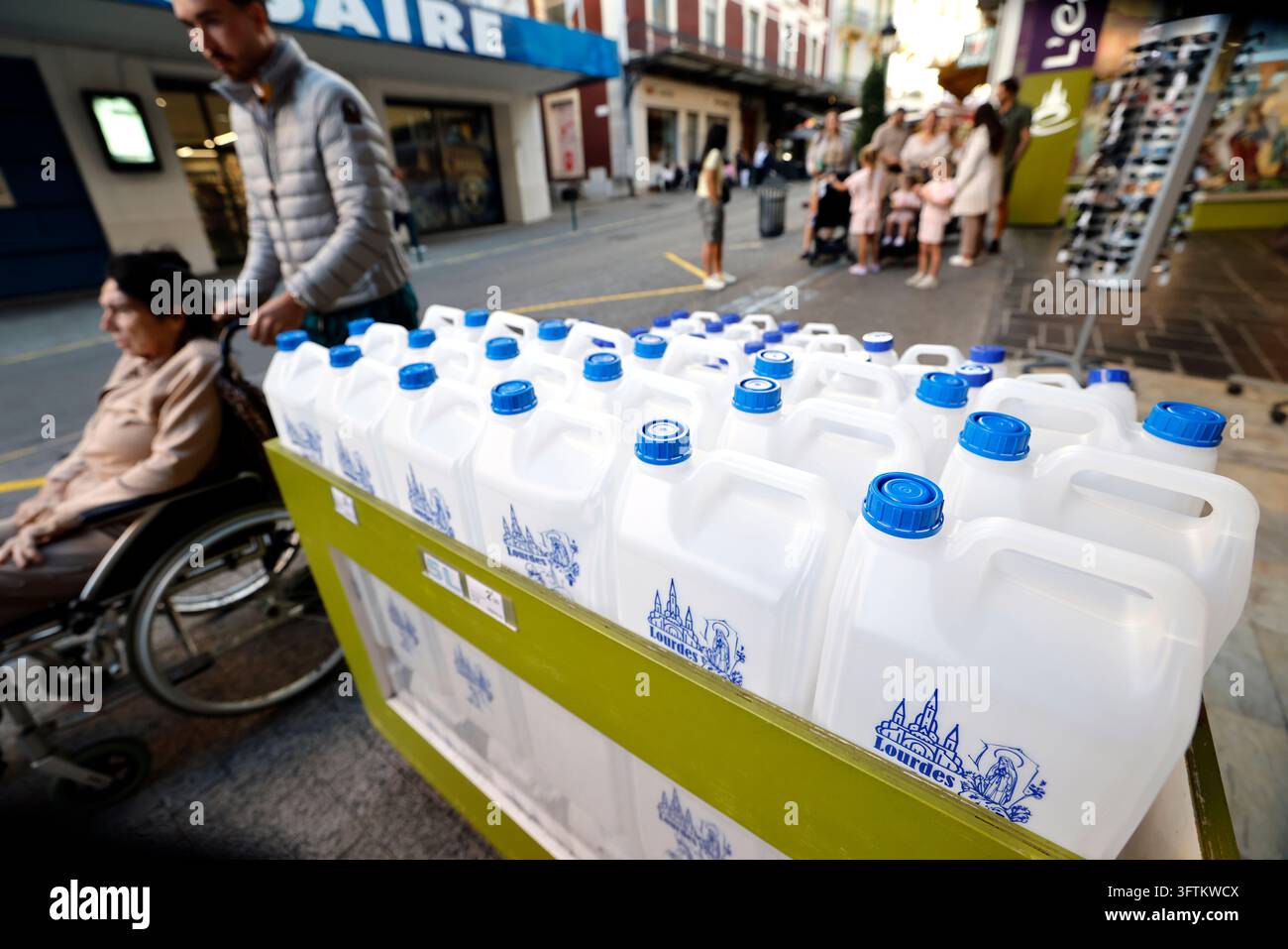 Weiße Plastikflaschen mit blauen Verschlüssen sind vor einem Souvenirladen aufgereiht, während Menschen in Rollstühlen vorbeifahren. Lourdes, Frankreich. Stockfoto