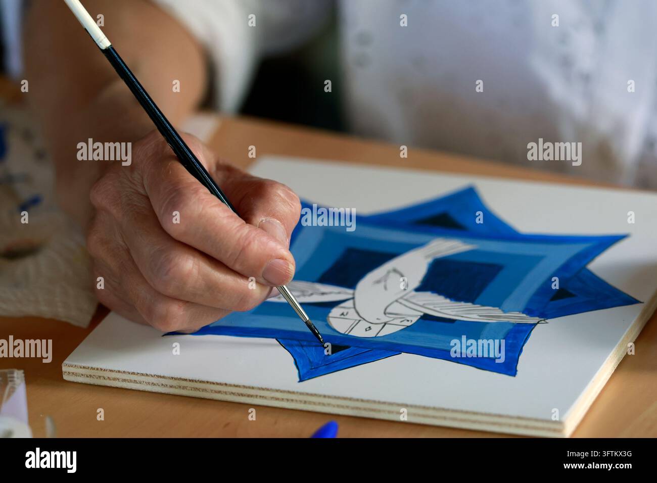 Ein Studierender Künstler trägt mit einem feinen Pinsel sorgfältig blaue Farbe auf eine Holztafel auf, die geometrische Muster aufweist, die den Heiligen Geist darstellen. Stockfoto