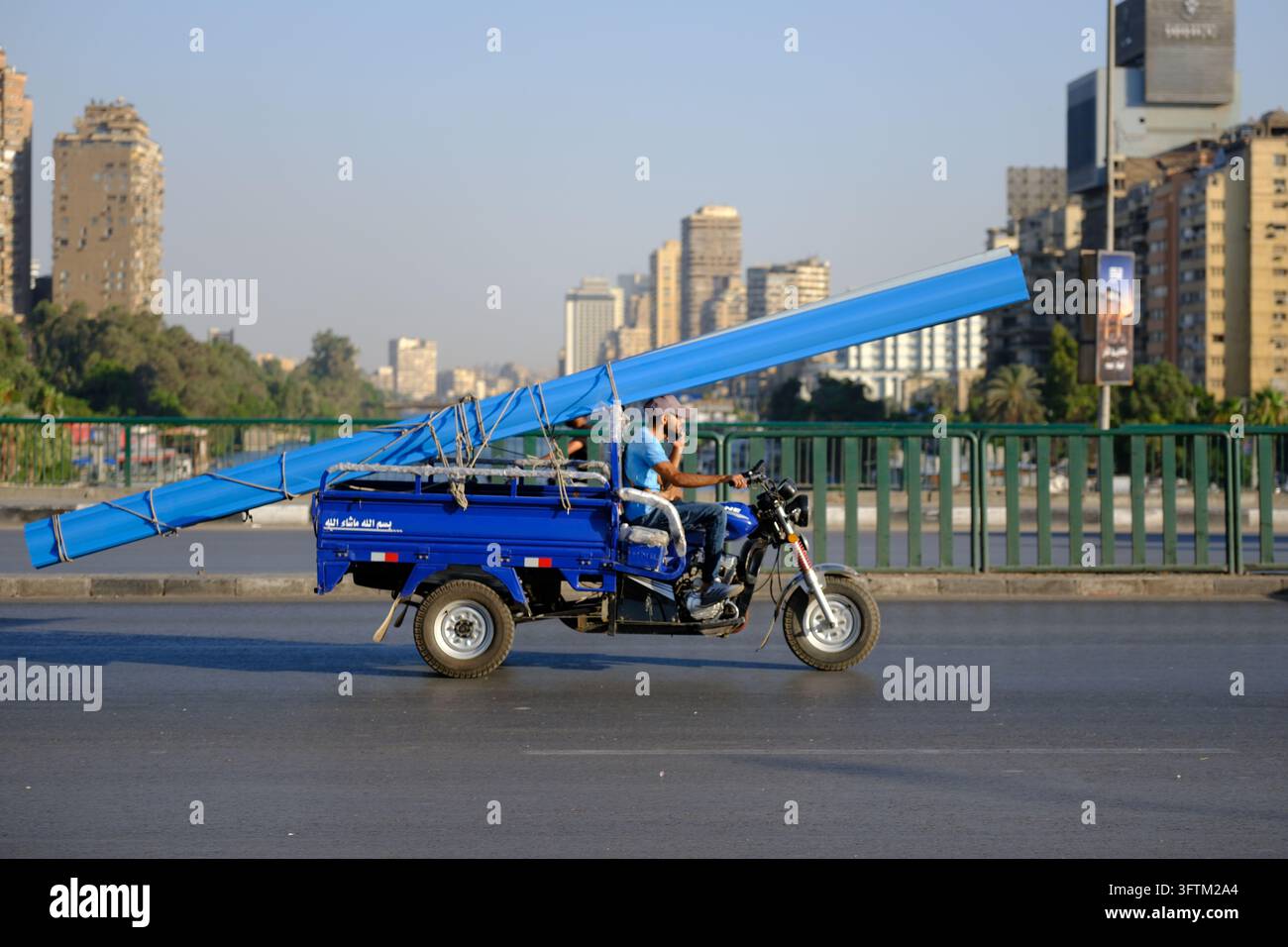 In Kairo, Ägypten, fährt ein Mann einen blauen dreirädrigen Karren mit langen Balken eine breite Allee hinunter. Stockfoto