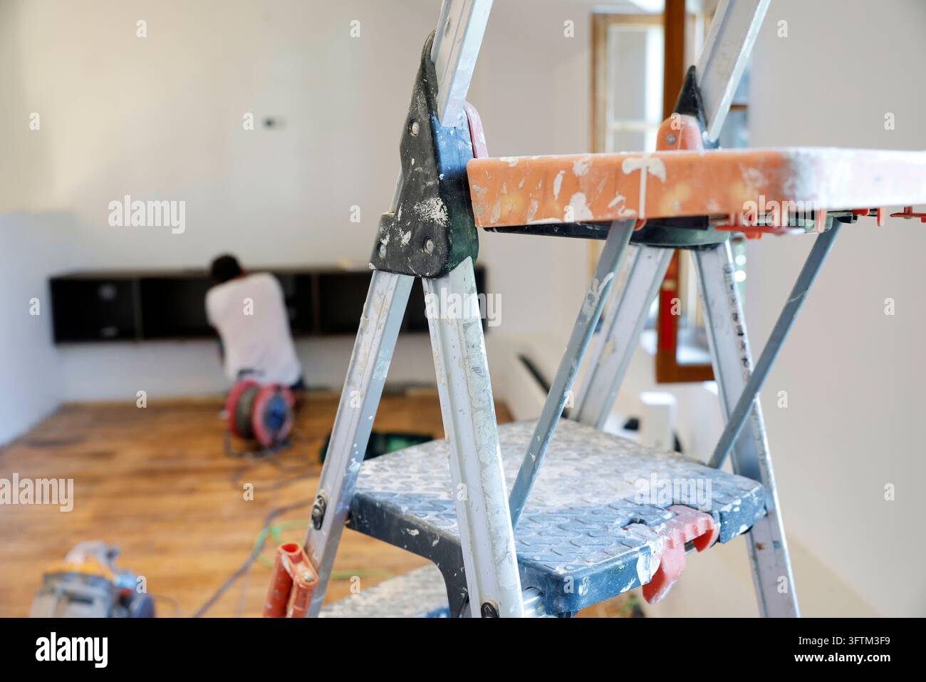 Eine Aluminium-Trittleiter steht im Vordergrund des renovierungsbedürftigen Raums, während ein Arbeiter im verschwommenen Hintergrund zu sehen ist. Stockfoto