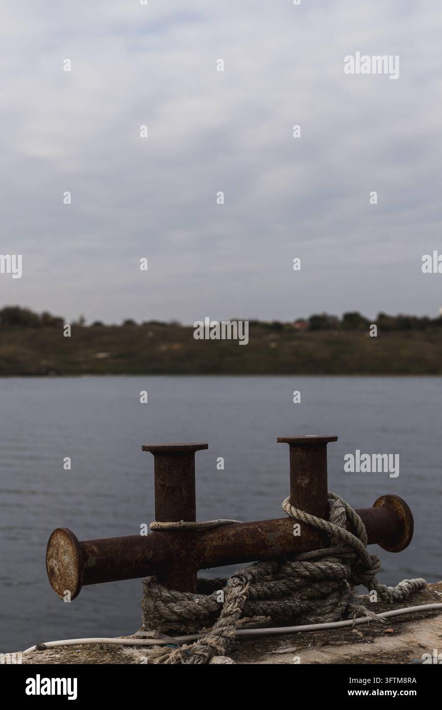 Rostiger Eisen-Pfahl mit gebundener Leine an einem Kai neben ruhigem Flusswasser unter einem bewölkten Himmel Stockfoto