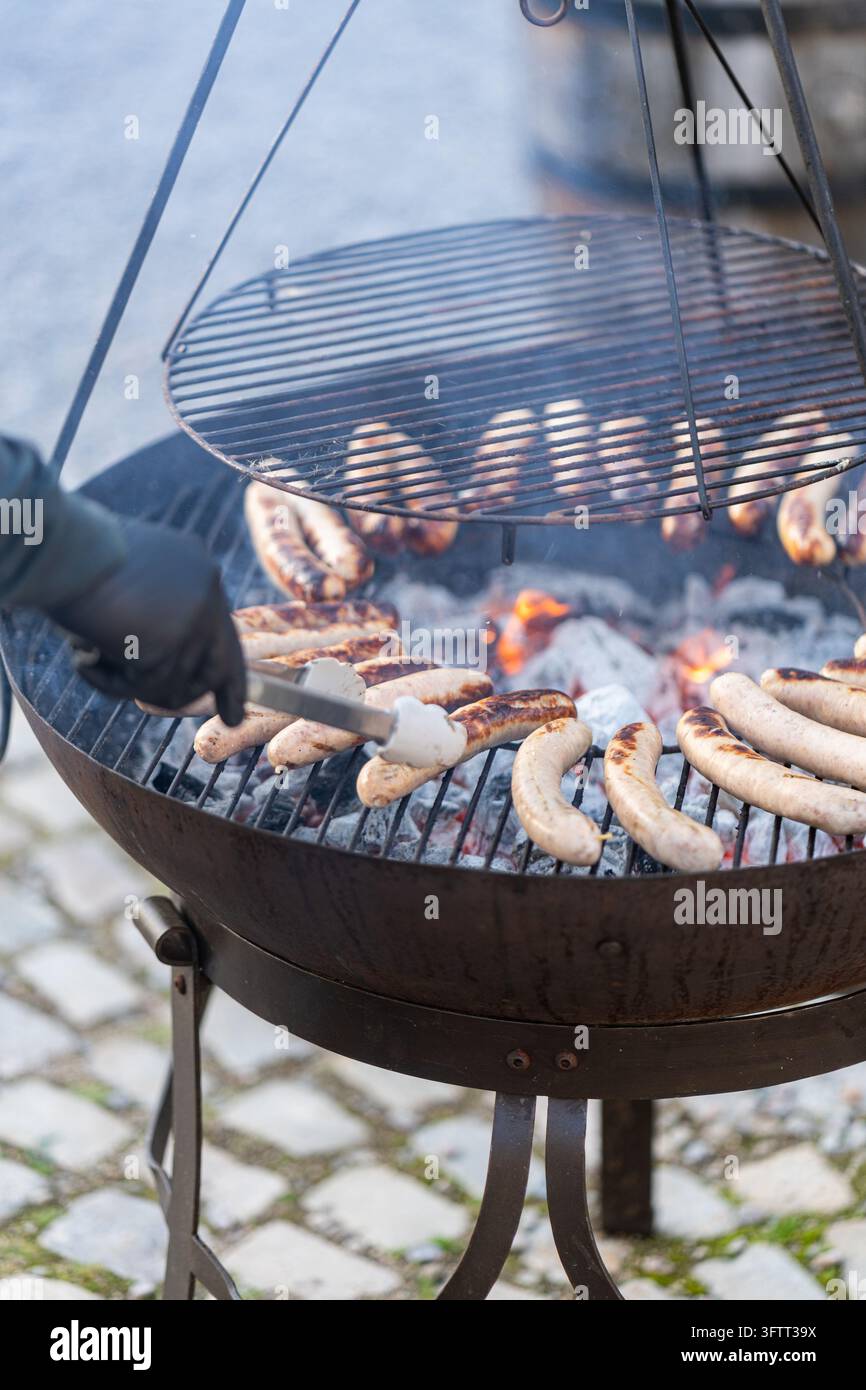 Würste, die über glühender Kohle auf einem Feuergrill mit hängendem Grill garen, werden mit einer Zange von einer behandschuhten Hand auf Kopfsteinpflaster gewendet Stockfoto