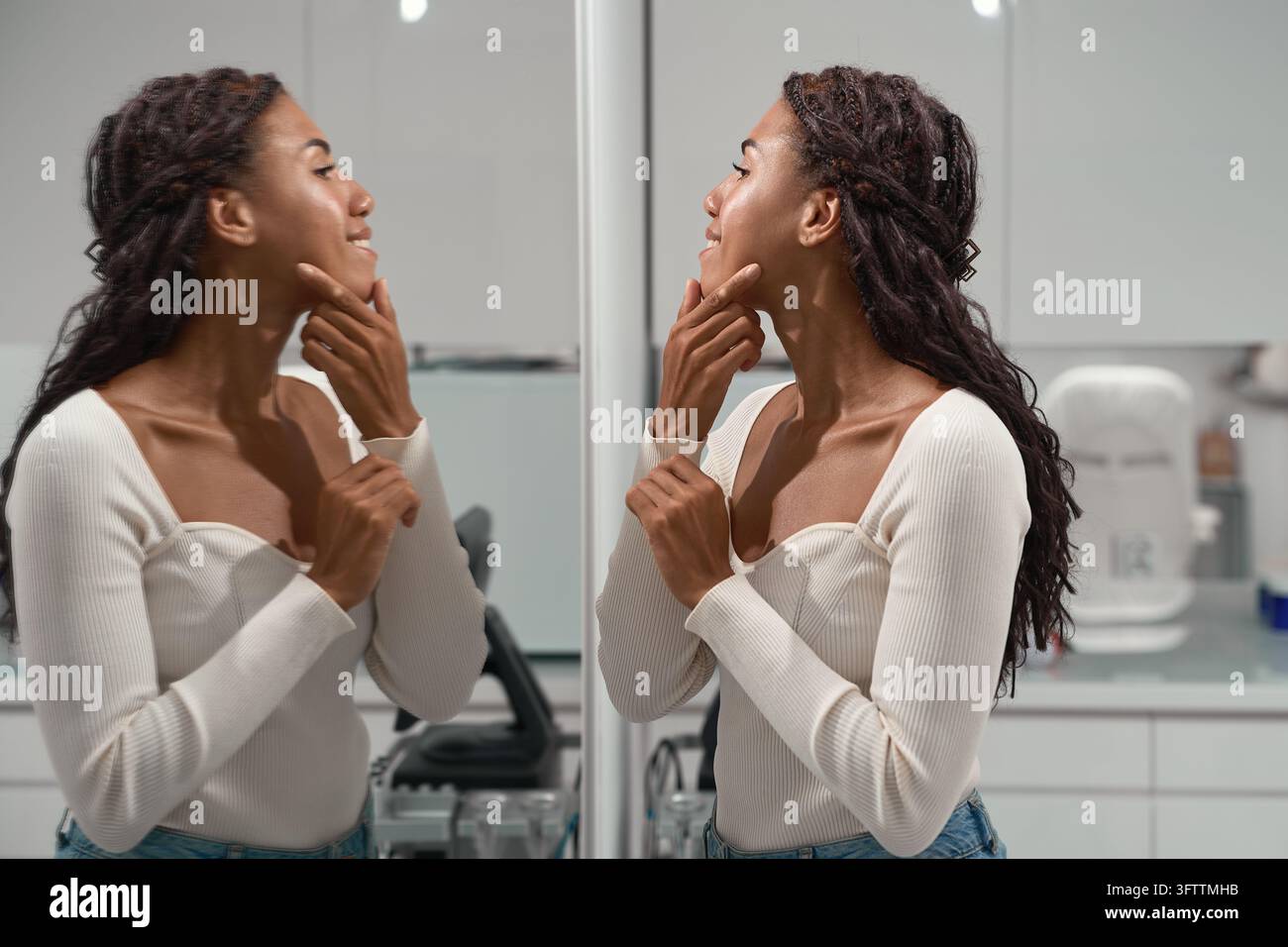 Afrikanische Frau lächelt in einen Spiegel, während sie mit einem Finger die Kieferlinie nachzieht und während einer Kosmetikberatung die Gesichts Konturen überprüft über Haut Straffung Stockfoto