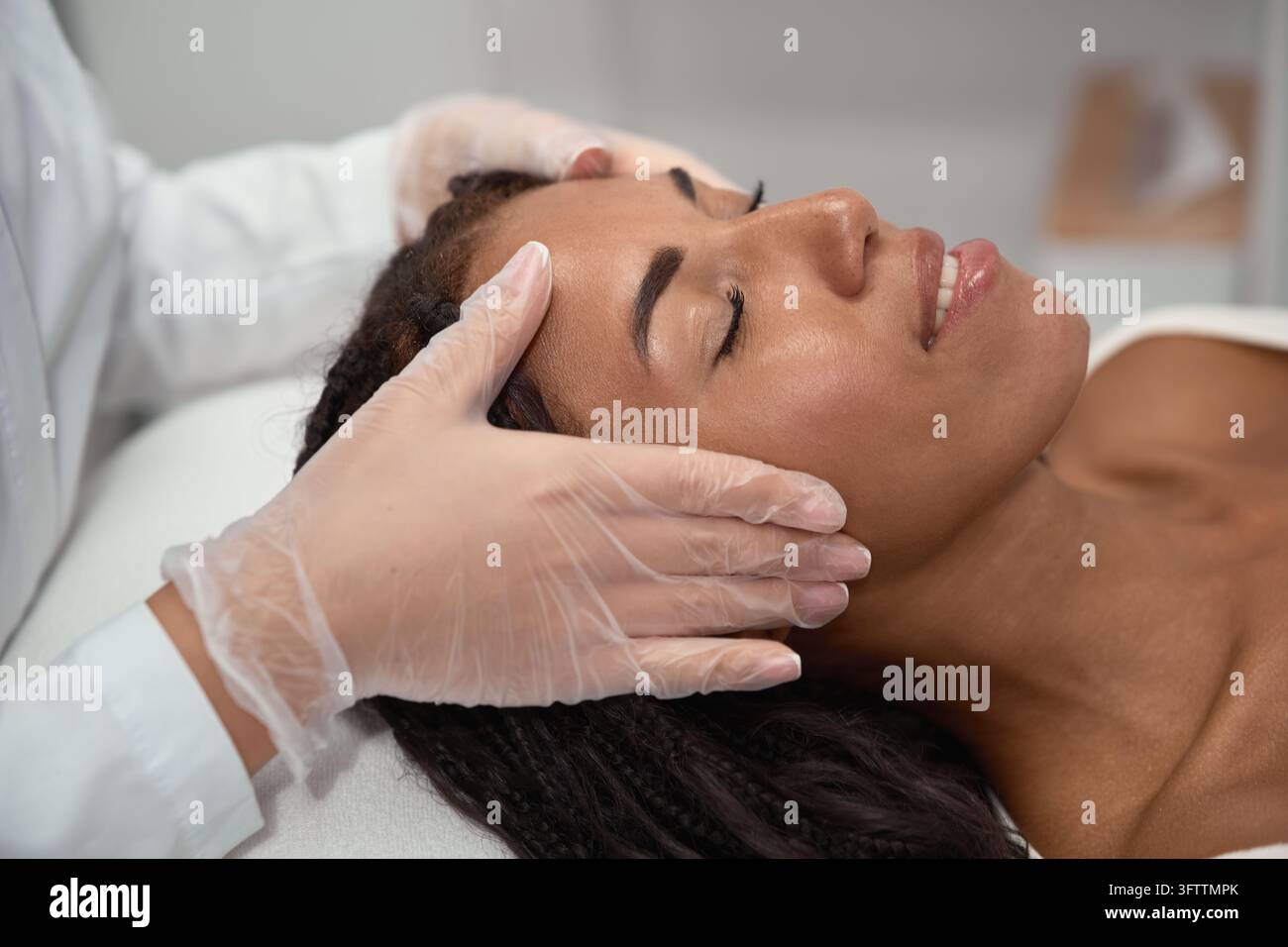 Mit kontrolliertem Fingerspitzen-Druck über den Brauen führt eine Kosmetikerin eine Gesichtsformungsmassage bei einer afrikanischen Frau durch, wobei sie sich auf manuelles Lifting konzentriert Stockfoto