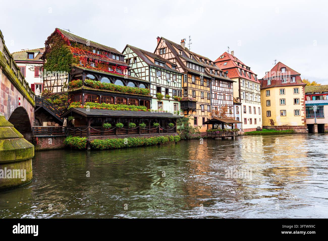 Farbenfrohe Fachwerkhäuser im Viertel Petite France von Straßburg, Frankreich, säumen einen ruhigen Fluss mit Spiegelungen im Wasser. Die traditionellen Gebäude zeigen Blumenkästen, von Efeu überwucherte Fassaden, eine Flussufer-Restaurantterrasse und eine nahe steinerne Brücke Stockfoto