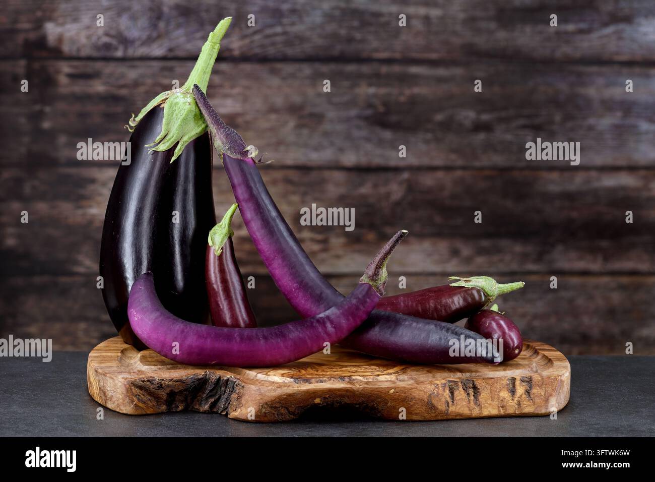 Verschiedene Sorten von frischen Auberginen, Solanum melongena, in verschiedenen Formen, Größen und Lila-Tönen, arrangiert auf einem rustikalen Holzschneidebrett. A Stockfoto