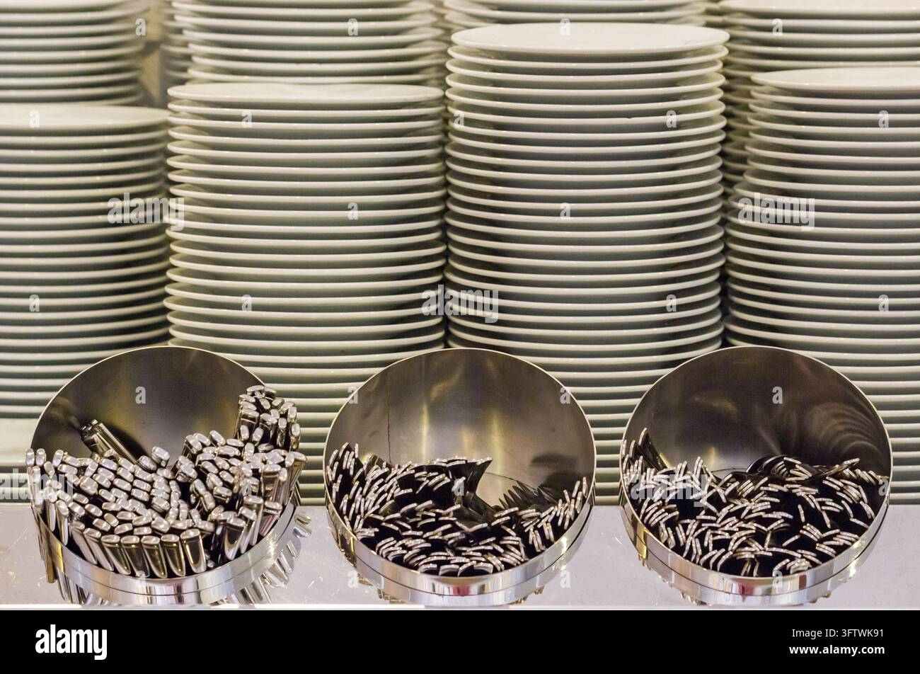 Ordentlich angeordnete Teller erheben sich in Stapeln hinter Metallhaltern, die mit Löffeln und Utensilien gefüllt sind, und schaffen eine organisierte Catering- oder Buffetszene Stockfoto