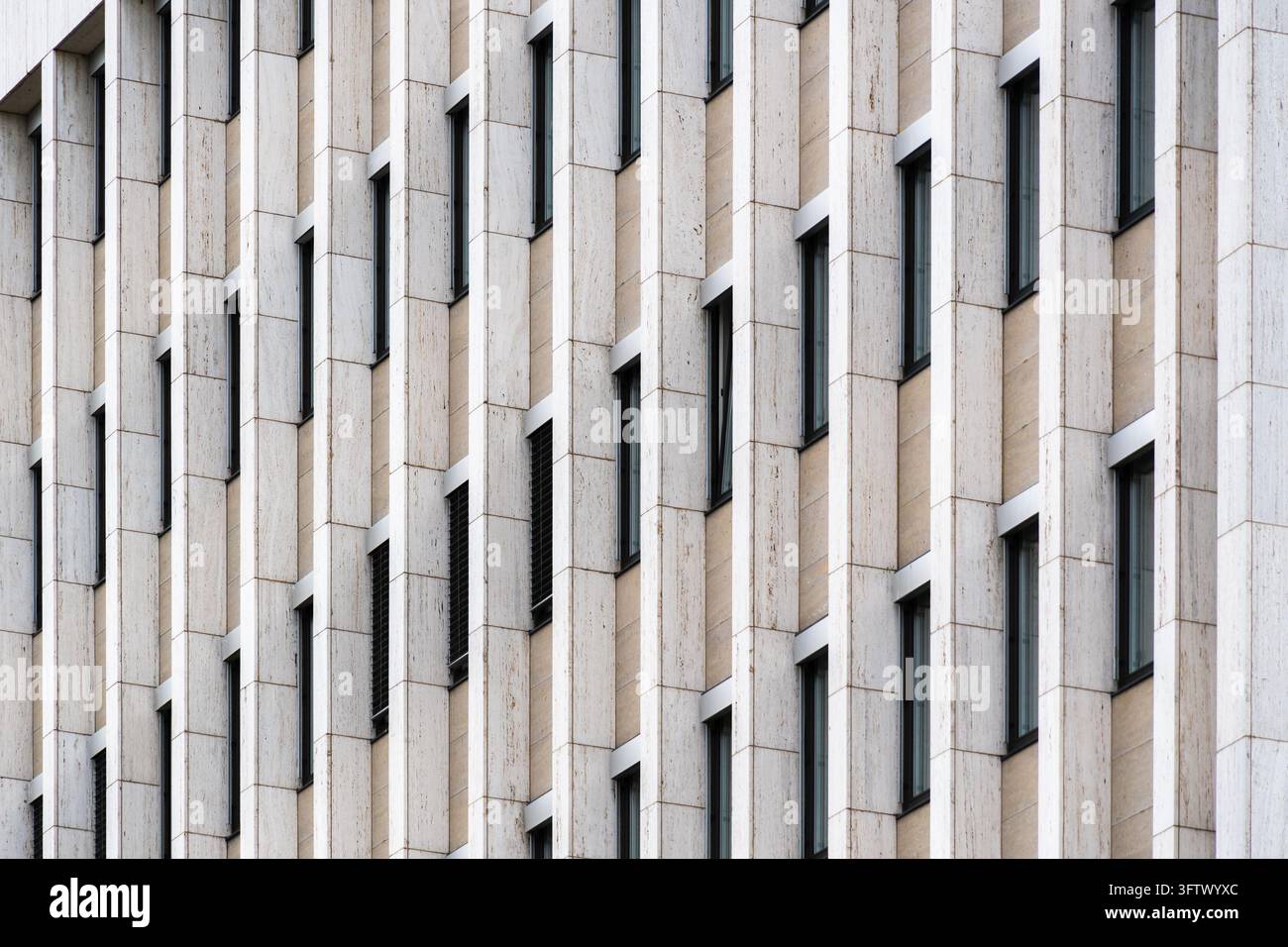 Schmale Fenster unterbrechen die helle Steinkladding eines Bürogebäudes. Wiederholte verticale Elemente und versetzte Fensterpositionen erzeugen eine geometrische Stockfoto