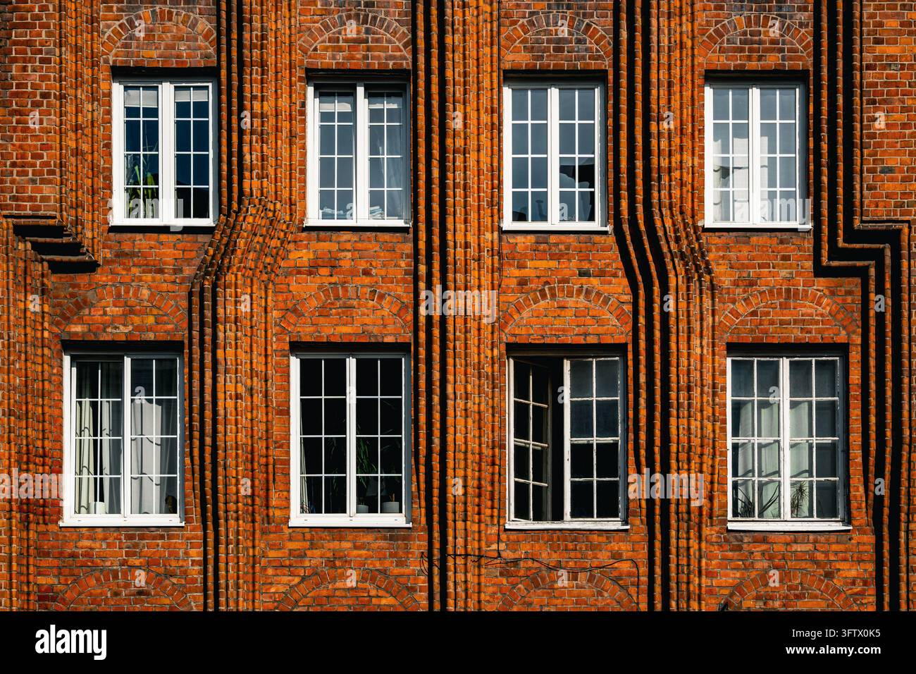 Weiß gerahmte Fenster unterbrechen eine rote Ziegelfassade, die mit flachen Mauerbögen und vorspringenden vertikalen Rippen verziert ist. Sonnenlicht betont die Textur Stockfoto