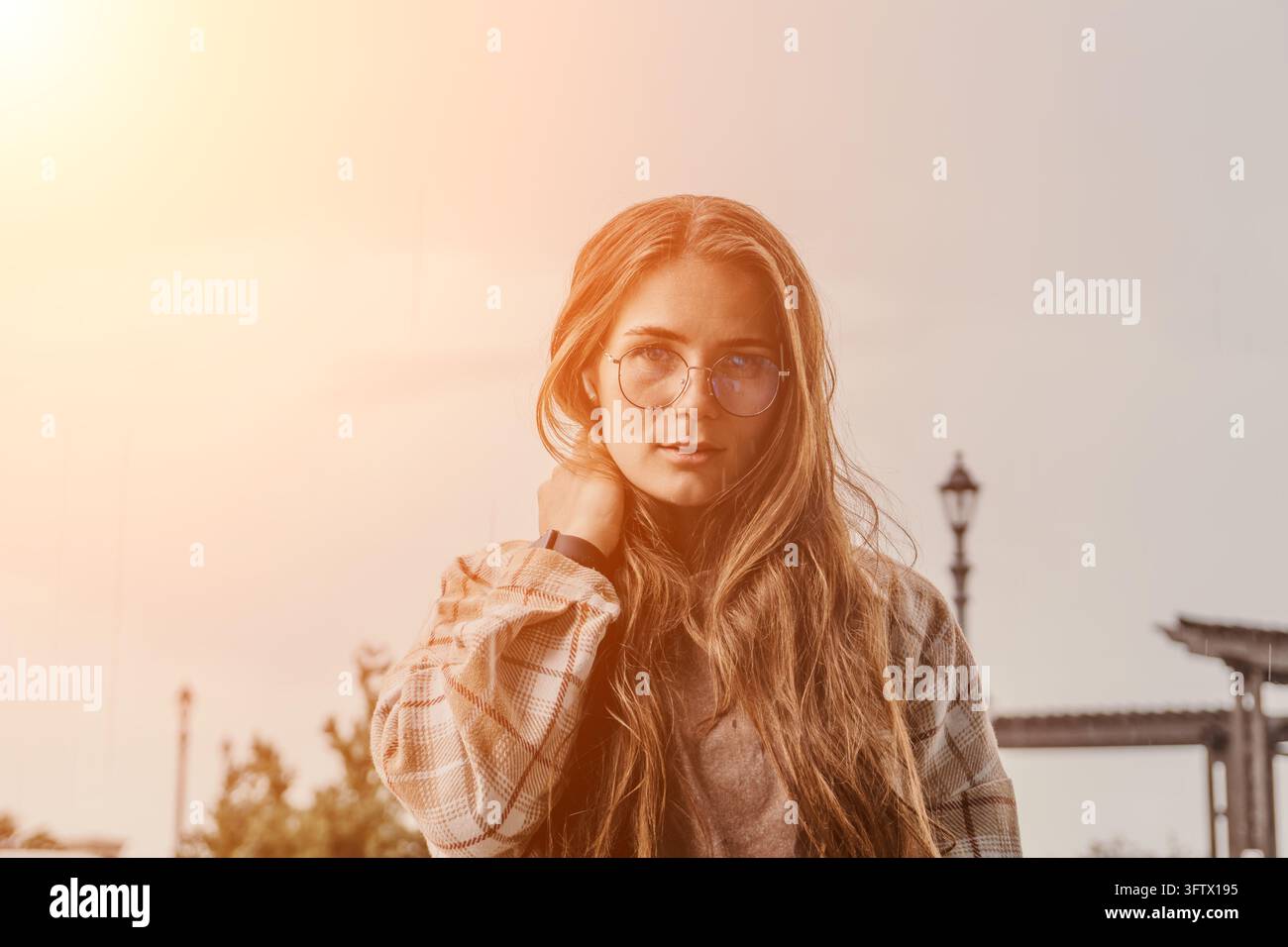 Frauenbrillenporträt einer jungen Frau mit langen Haaren, die eine karierte Jacke trägt, im Freien unter warmem Sonnenlicht beim Sonnenuntergang mit reichlich Kopierraum Stockfoto