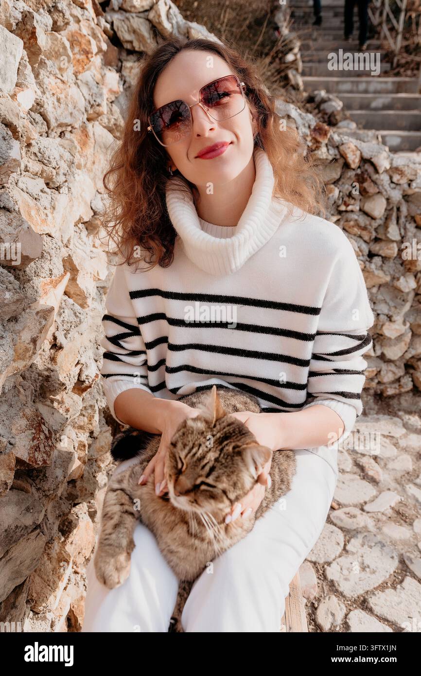 Frau mit Katzensonnenbrille sitzt an einer Steinmauer und streichelt eine freundliche getigerte Katze im warmen Herbstsonnenschein Stockfoto