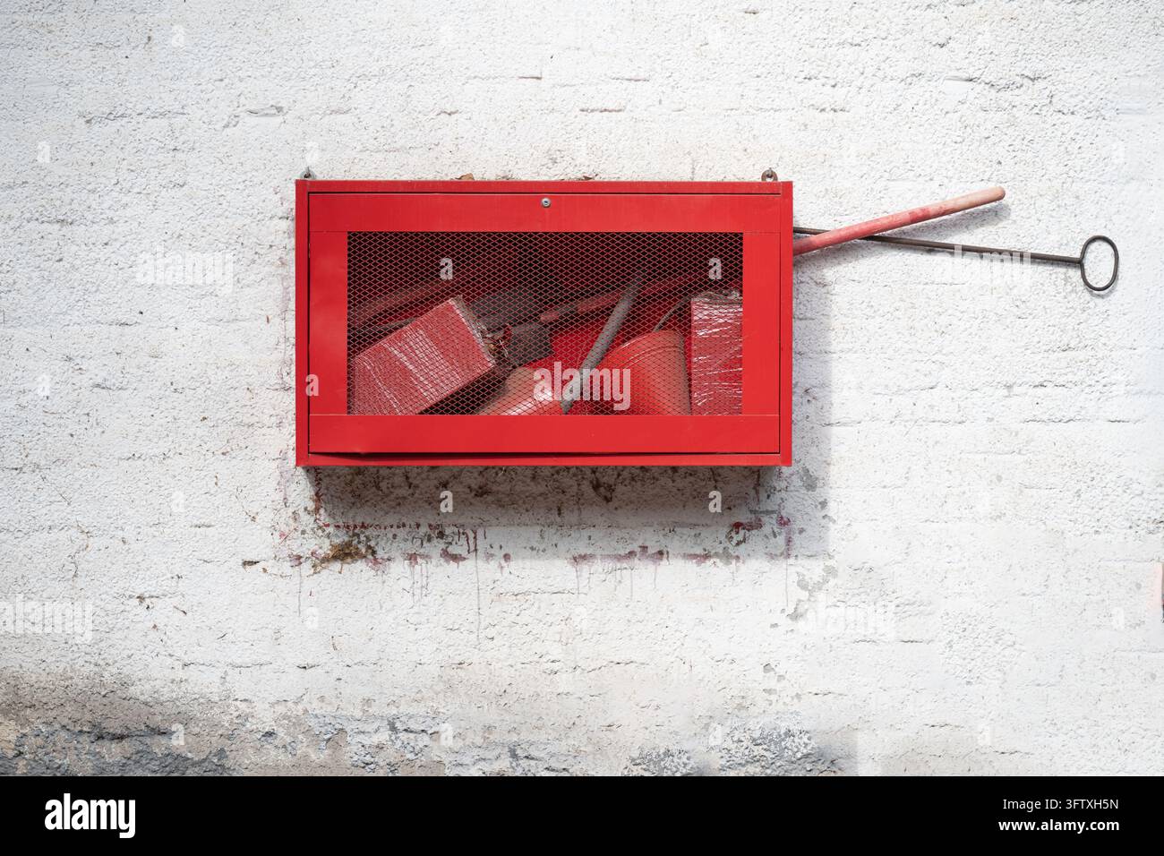 Roter Brandschutzschrank mit Notfallausrüstung an einer weißen strukturierten Wand. Industrie-Brandschutzbox mit Werkzeugen, Axt und Schaufel. Stockfoto