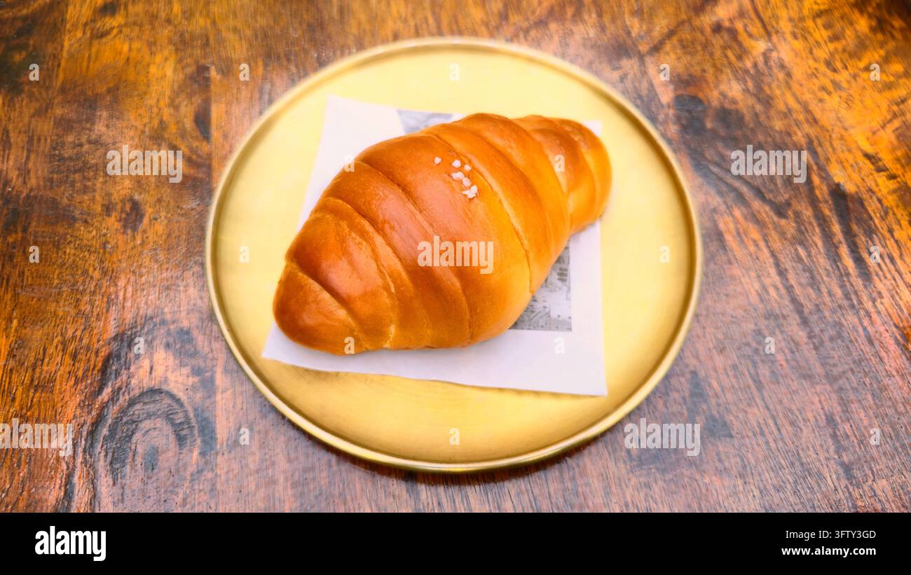 Japanisches Salzbrot mit einer glänzenden goldenen Kruste, serviert auf einem Teller mit einer Gabel auf einem rustikalen Holztisch, ein beliebtes weiches, buttriges Backwarenbrot und Café-Snack - Smartphone-aufgenommenes Stockfoto
