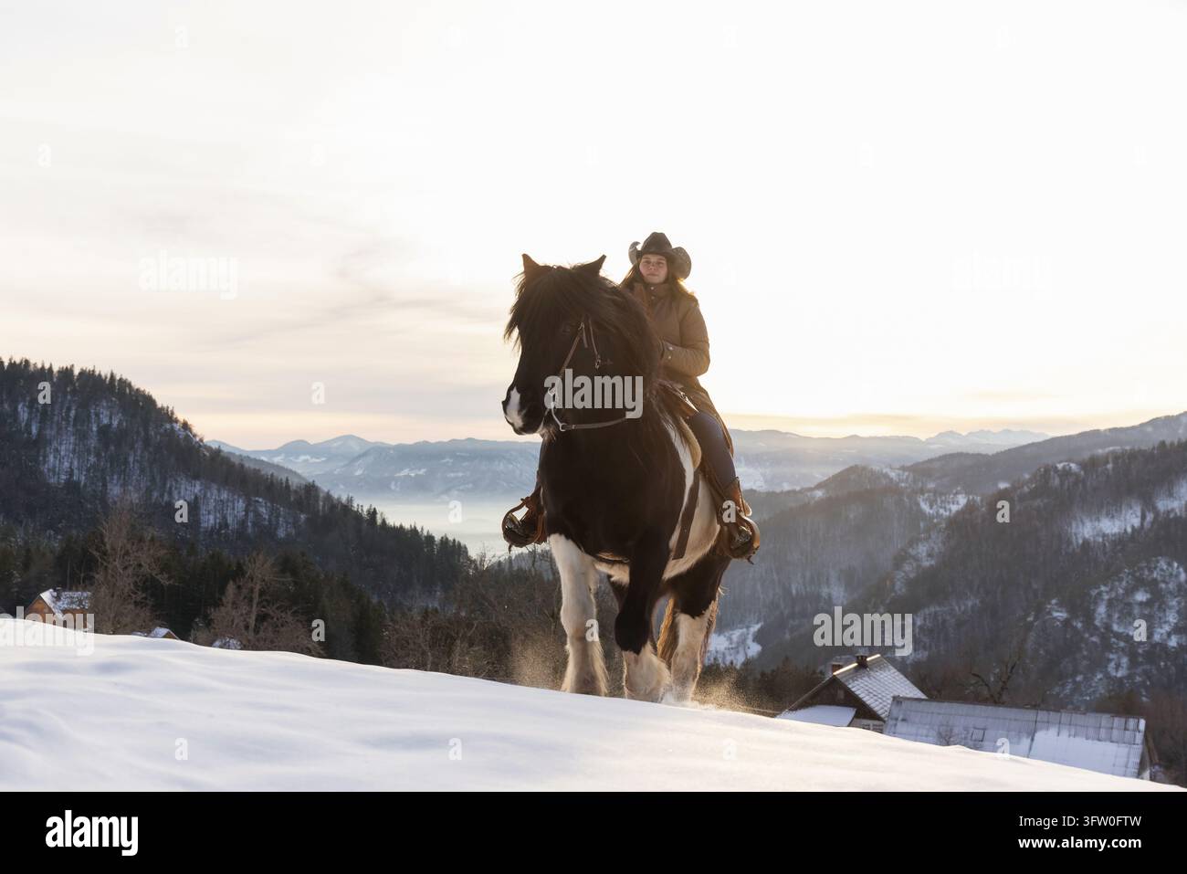 Frau genießt die Zeit im Freien während eines Winterausflugs, reitet im Goldenen Licht durch den Schnee in den Bergen auf einem Pferd. Stockfoto
