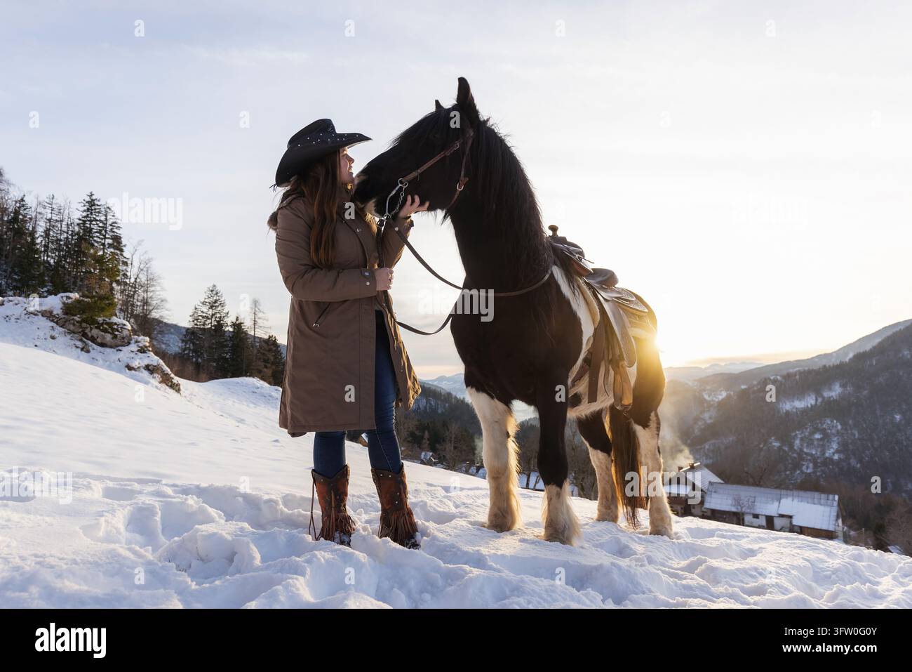 Frau verbringt einen friedlichen Moment mit ihrem Pferd in den schneebedeckten Bergen und genießt ihre Gesellschaft inmitten der Winterlandschaft. Stockfoto