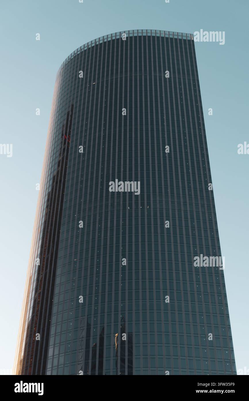 Eine niedrig angelegte Ansicht eines hohen Wolkenkratzers mit einer gebogenen dunklen Glasfassade. Dünne vertikale Linien teilen die Fenster. Reflexionen anderer Strukturen sind sichtbar Stockfoto