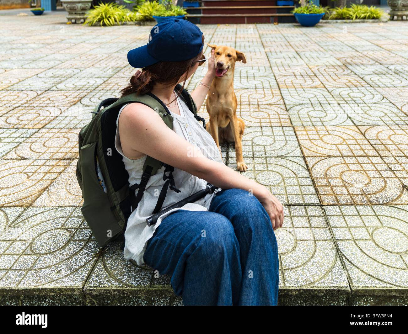Frau mit blauer Mütze sitzt auf gefliesten Stufen und streichelt einen goldbraunen Hund, Con Dao, Vietnam Stockfoto