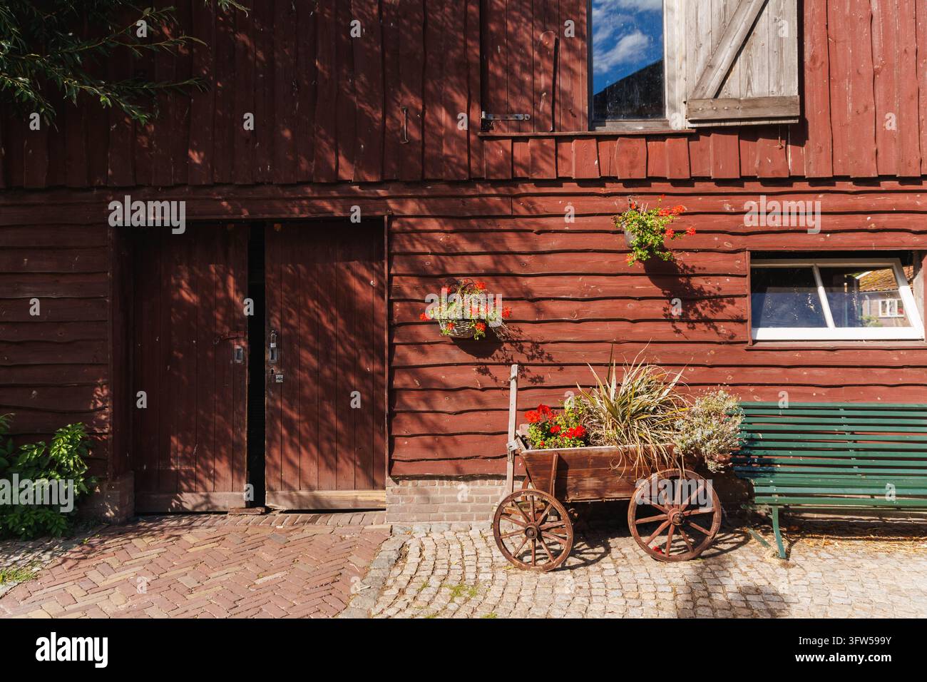 Sonnenbeschienene rote Holzscheune mit Brettertüren, Geranienblumenkarre und grüner Bank auf einem gepflasterten Hof Stockfoto