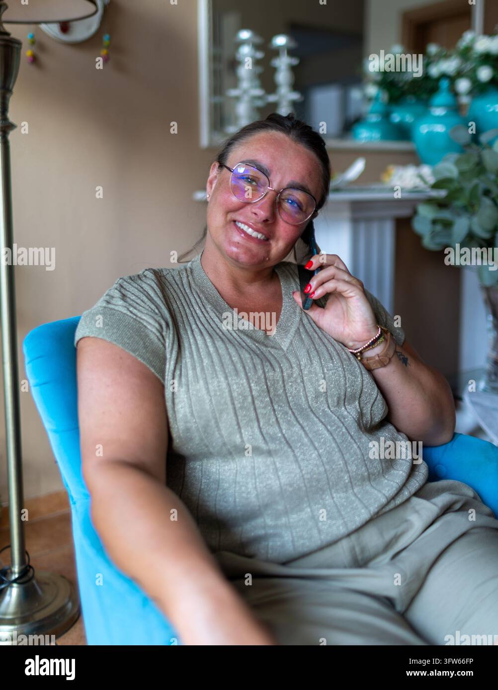Lächelnde Frau schaut in die Kamera, während sie von einem bequemen Sessel zu Hause aus telefoniert. Stockfoto