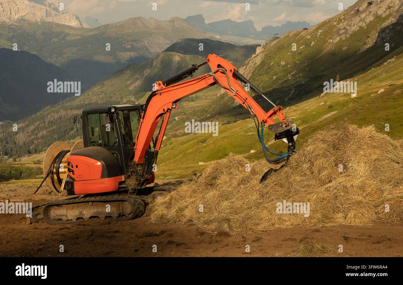 Orange Mini-Bagger mit Kettenlaufwerk arbeitet an einem steilen Alpenhang in den Dolomiten, Norditalien, mit Baumaterialien Stockfoto