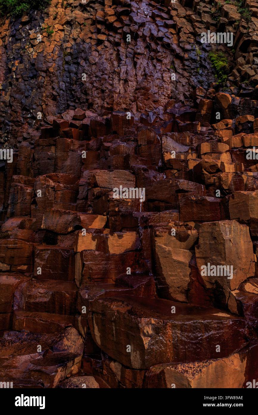 Eine detaillierte vertikale Aufnahme von strukturierten hexagonalen Basaltsteinblöcken, die dunkle nasse Oberflächen und Flecken von leuchtend orangefarbenem Rostmineral zeigen. Stockfoto