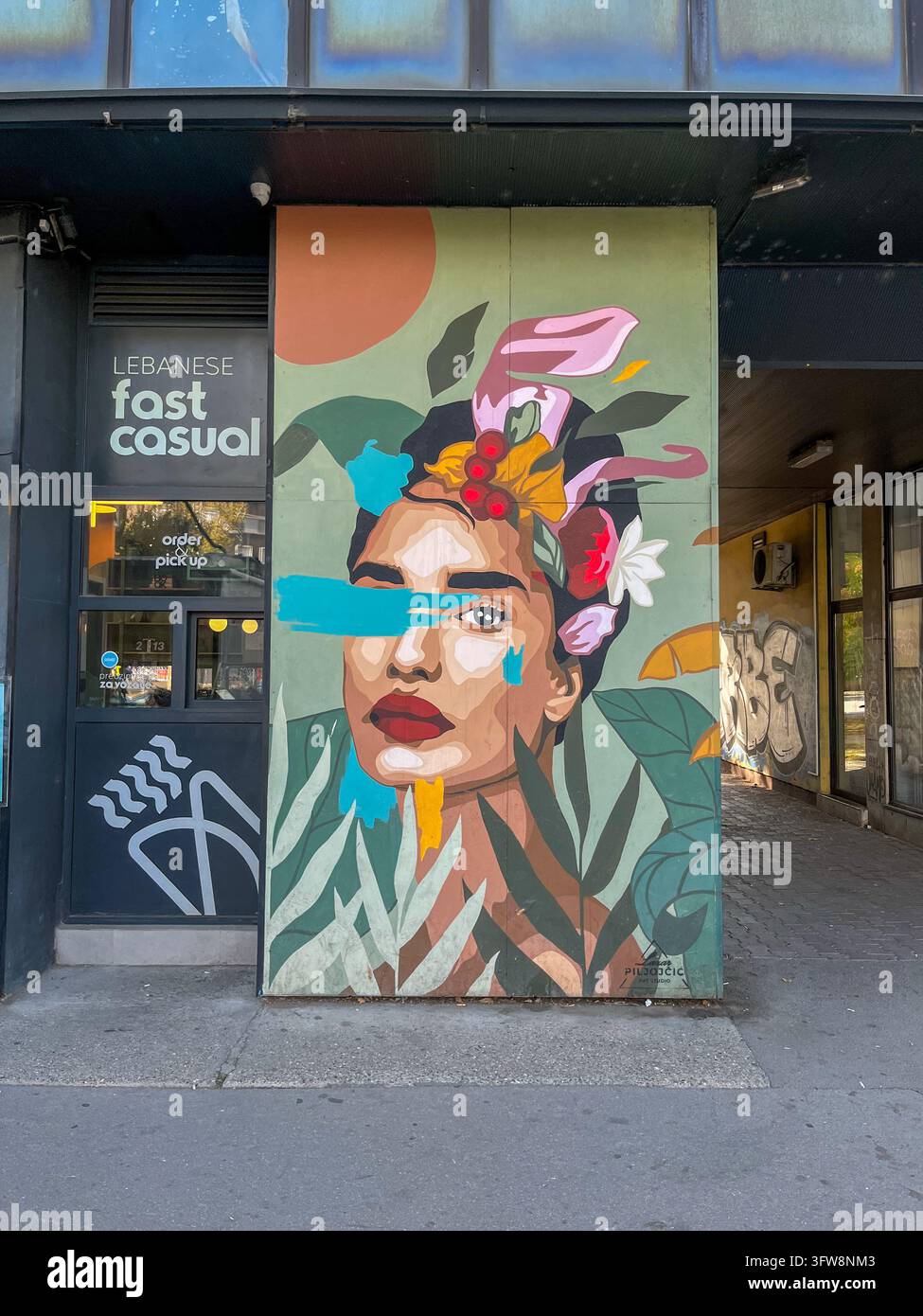 Novi Sad, Serbien - 18. Oktober 2026: Farbige Straßenkunst-Wandmalerei, die ein stilisiertes Frauenporträt zeigt, umgeben von tropischen Blättern und Blumen - Smartphone-aufgenommenes Stockfoto