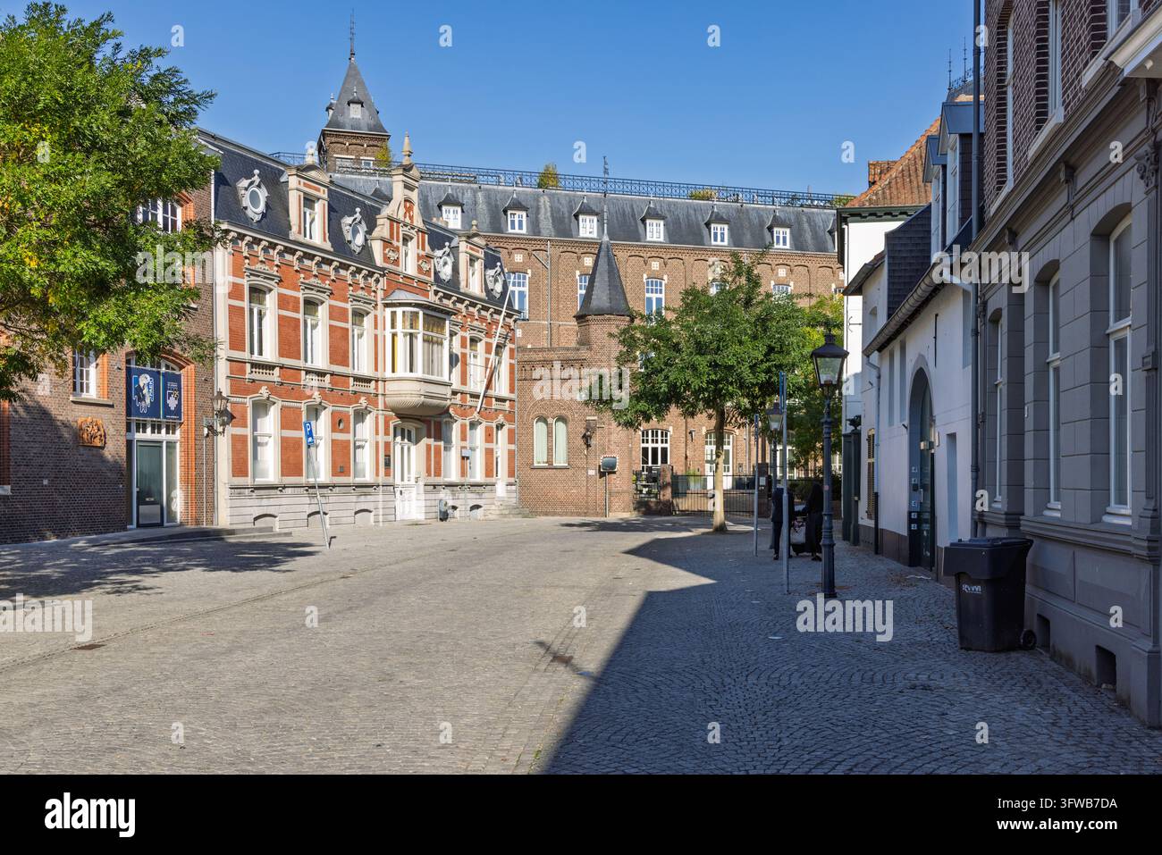 Sittard, Niederlande: Ruhiger gepflasterter Platz Kloosterplein mit historischen roten Backsteingebäuden, traditionellen Dachgauben und üppigen grünen Bäumen unter einem Bri Stockfoto