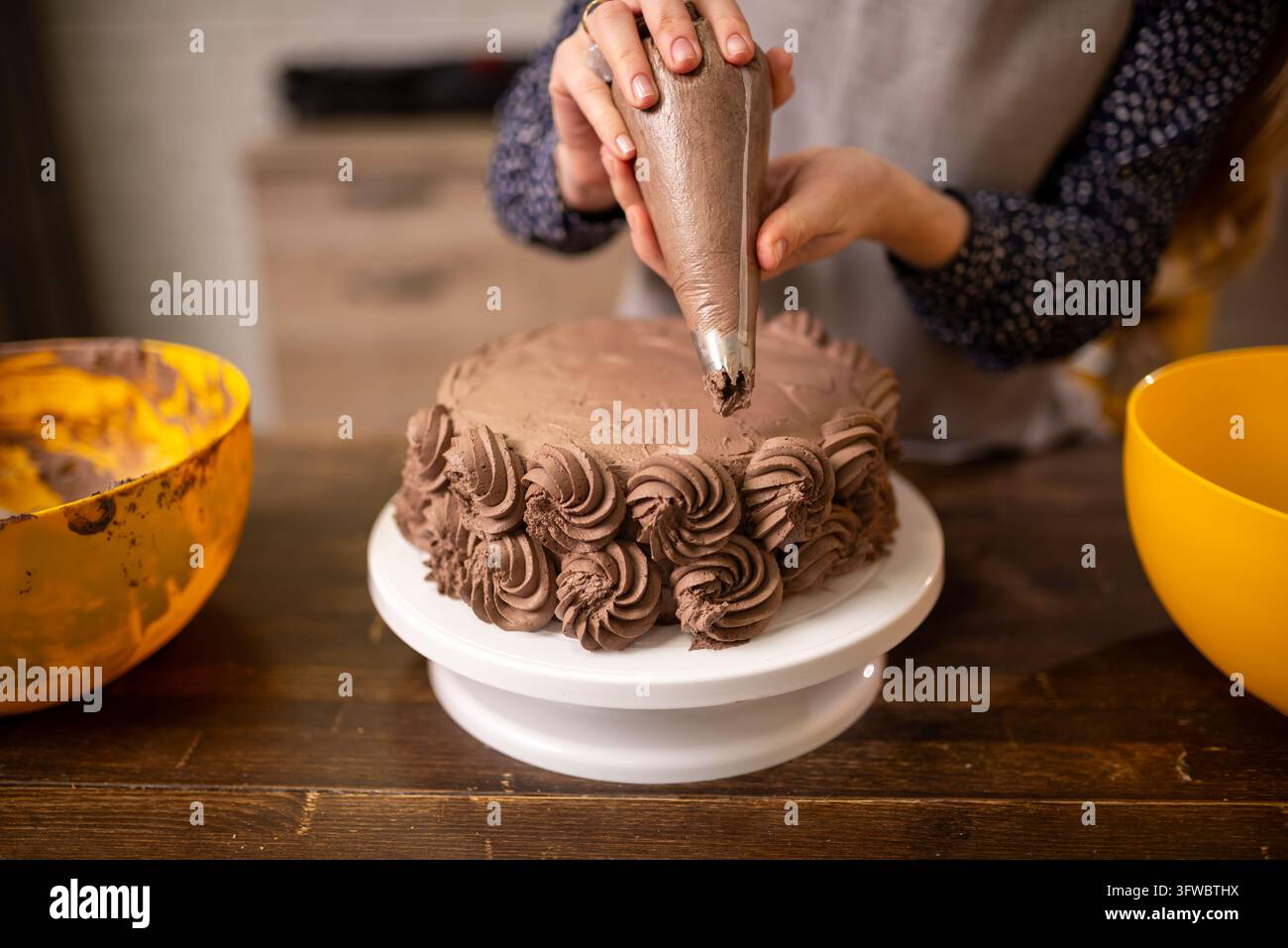 Nahaufnahme von Händen, die Schokoladen-Buttercreme-Rosetten auf einem runden Schichtkuchen in einer gemütlichen Kücheneinrichtung spritzen Stockfoto