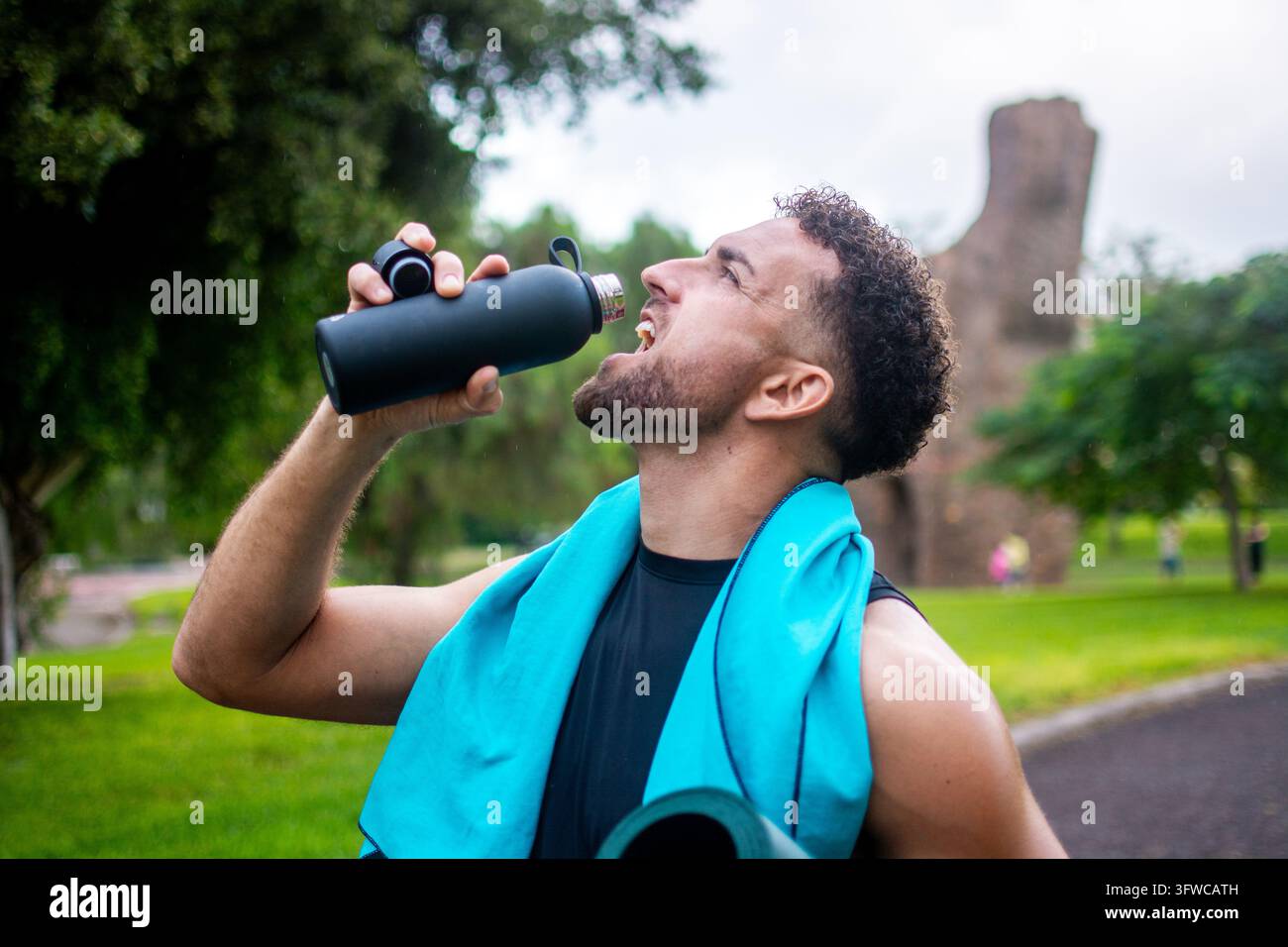 Seitenansicht eines muskulösen Mannes mit türkisfarbenem Handtuch um den Hals, der nach dem Sport in einem Fitnessstudio Wasser aus einer schwarzen wiederverwendbaren Sportflasche trinkt Stockfoto