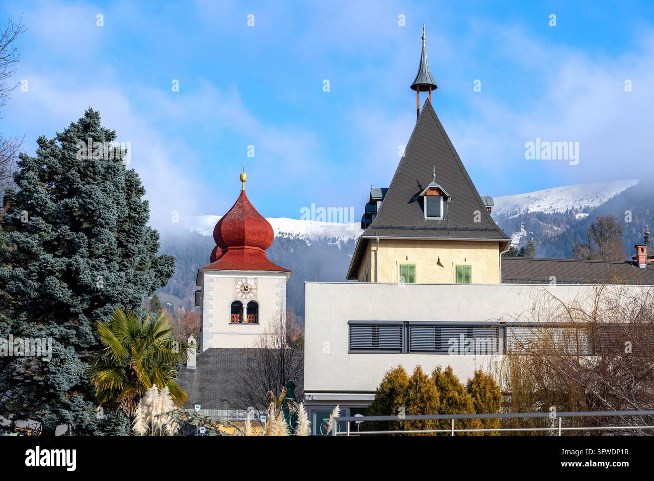 Historischer europäischer Kirchturm mit einer roten Zwiebelkuppel neben traditioneller spitzdachiger Architektur, umgeben von immergrünen Bäumen und schneebedeckten Bergen Stockfoto