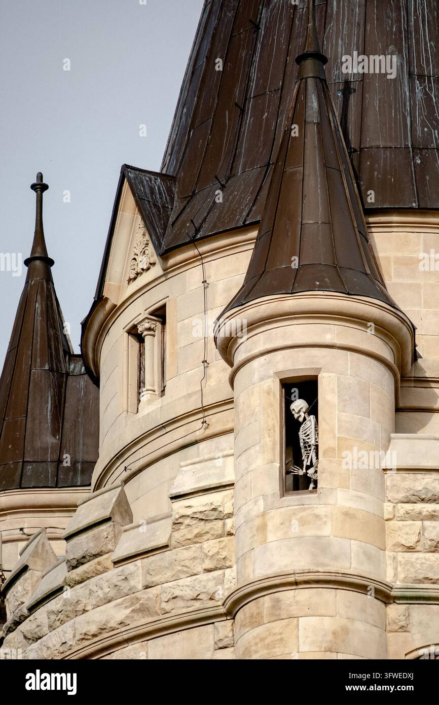 Steinerner Burgturm mit kegelförmigen Kupferdächern und Skelettfigur in schmalem Gewölbefenster, beiger Mauerwerk und dekorativer Steinmetzarbeit Stockfoto