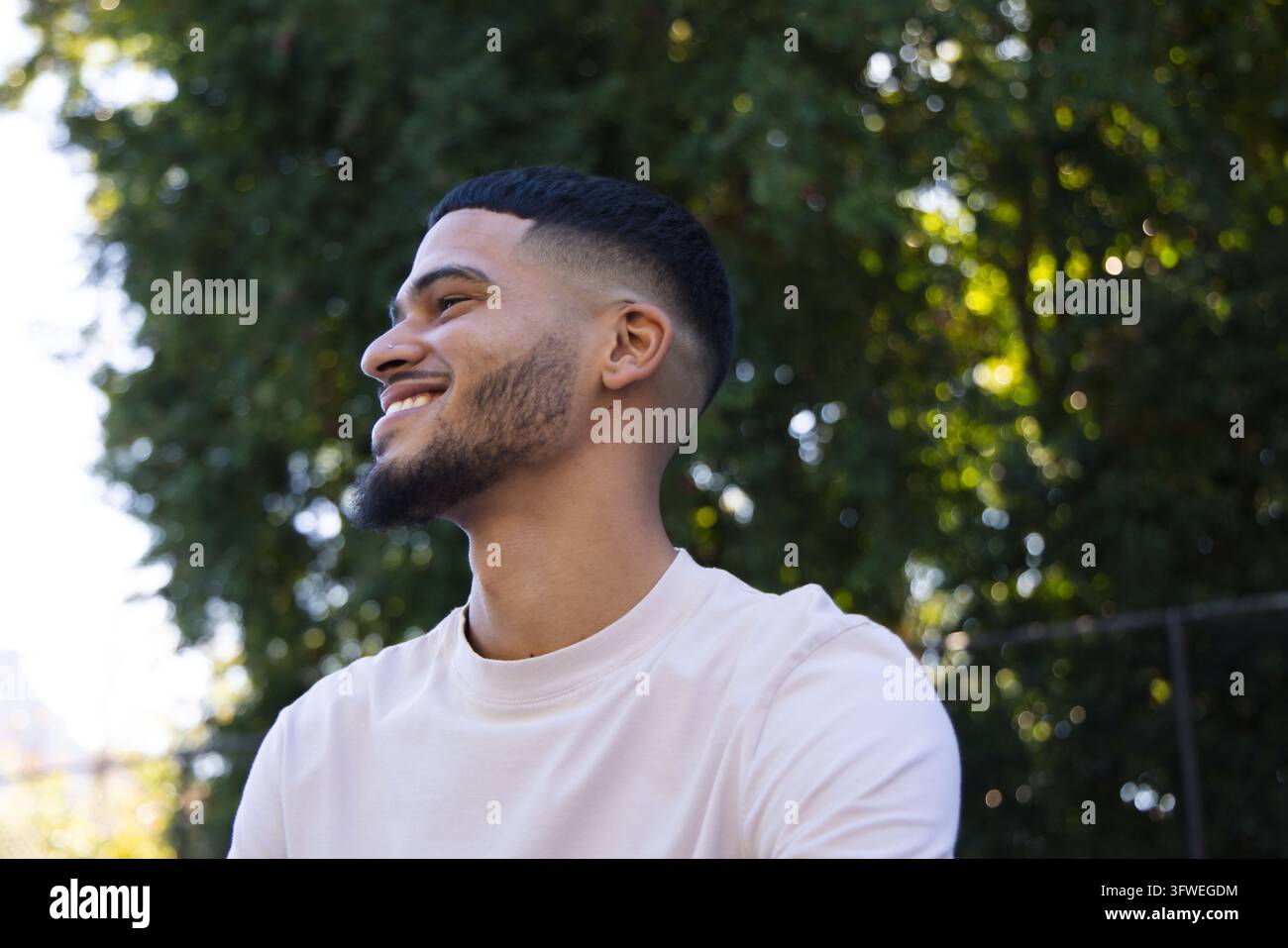Afroamerikaner lächelt, schaut nach links, trägt ein helles Crew-Neck-T-Shirt im Park in der Nähe eines Metallzauns Stockfoto