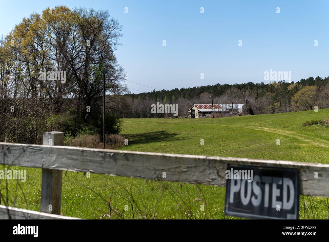 Blick auf eine ländliche Landschaft mit einem weißen Holzzaun, einem Hinweisschild, einem grünen Feld und einer rustikalen Scheune, die zwischen Bäumen unter einem klaren blauen Himmel in Mar liegt Stockfoto