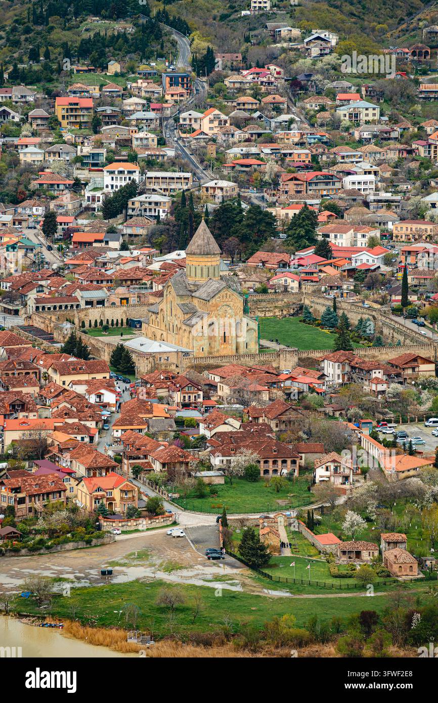 Luftbild der Svetitskhoveli-Kathedrale, umgeben von terracottafarbenen Dächern von Mtskheta, Georgien, neben einem schlammigen Fluss mit Frühlingsblüten und grünem Laich Stockfoto