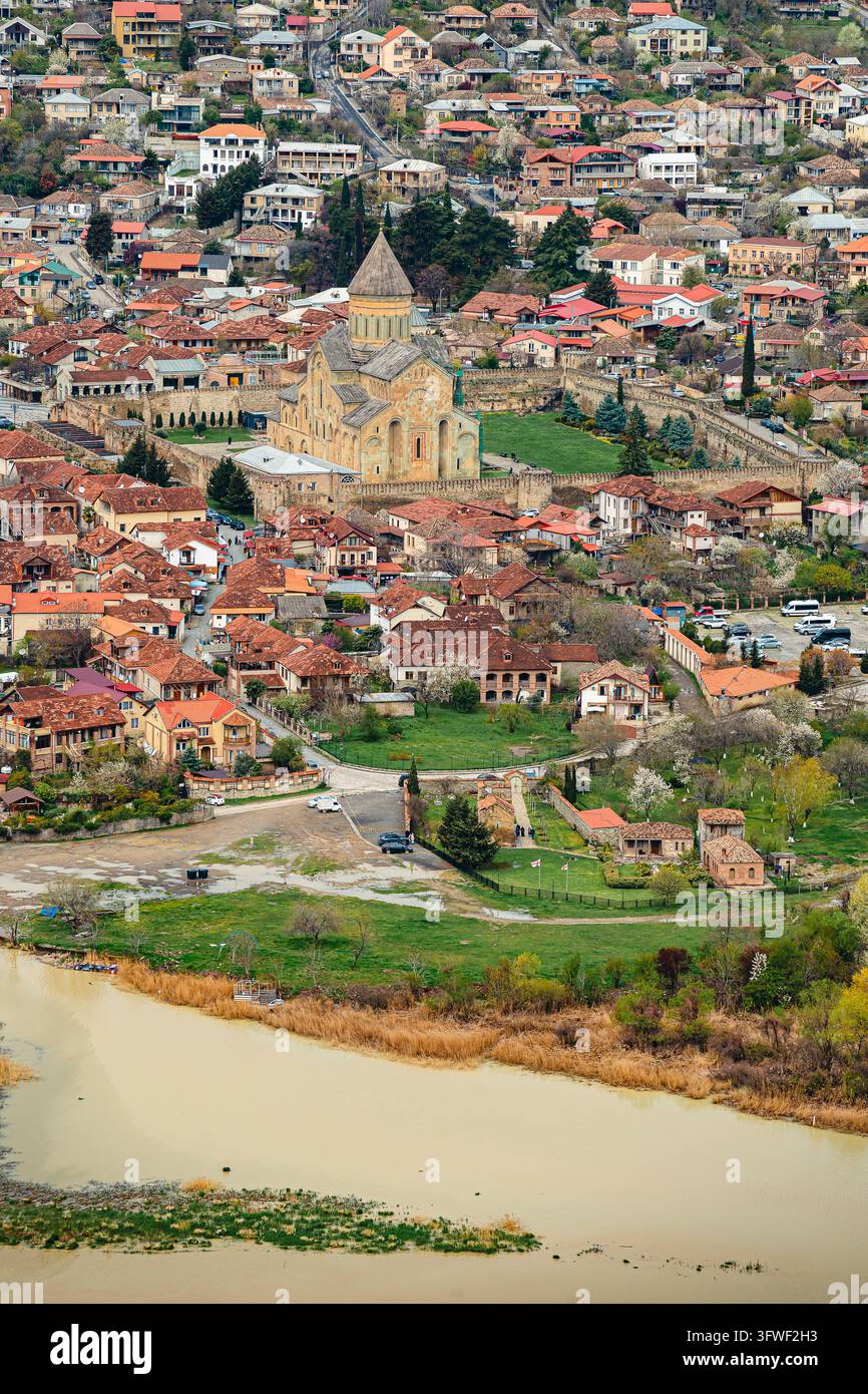 Luftbild der Svetitskhoveli-Kathedrale, umgeben von terracottafarbenen Dächern von Mtskheta, Georgien, neben einem schlammigen Fluss mit Frühlingsblüten und grünem Laich Stockfoto