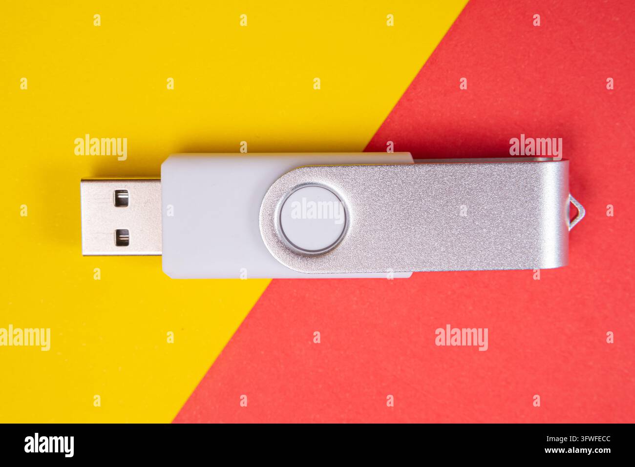 Ein moderner silberner und weißer USB-Speicherstick mit Drehfunktion liegt auf einem lebendigen, geteilten Hintergrund aus Gelb und Rot. Dieses Bild betont Speicher und Technik Stockfoto