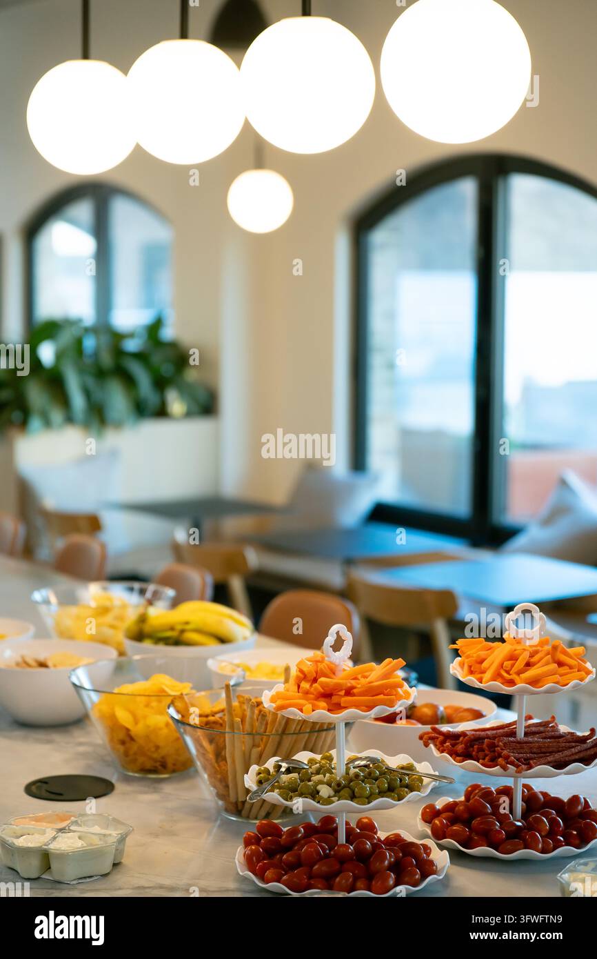 Ansicht eines Snack-Buffets mit Schalen voller Chips, Bananen, Karottenstiften, Oliven und Kirschtomaten auf einem Tisch in einem modernen Büro mit Pendelleuchten. L Stockfoto