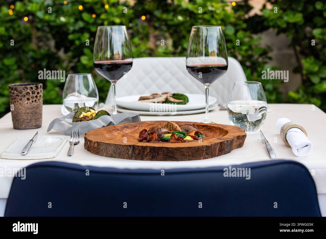 Ein elegant gedeckter Außentisch zeigt Weingläser, Teller mit Speisen und Besteck vor einem Hintergrund aus grünem Laub. Stockfoto