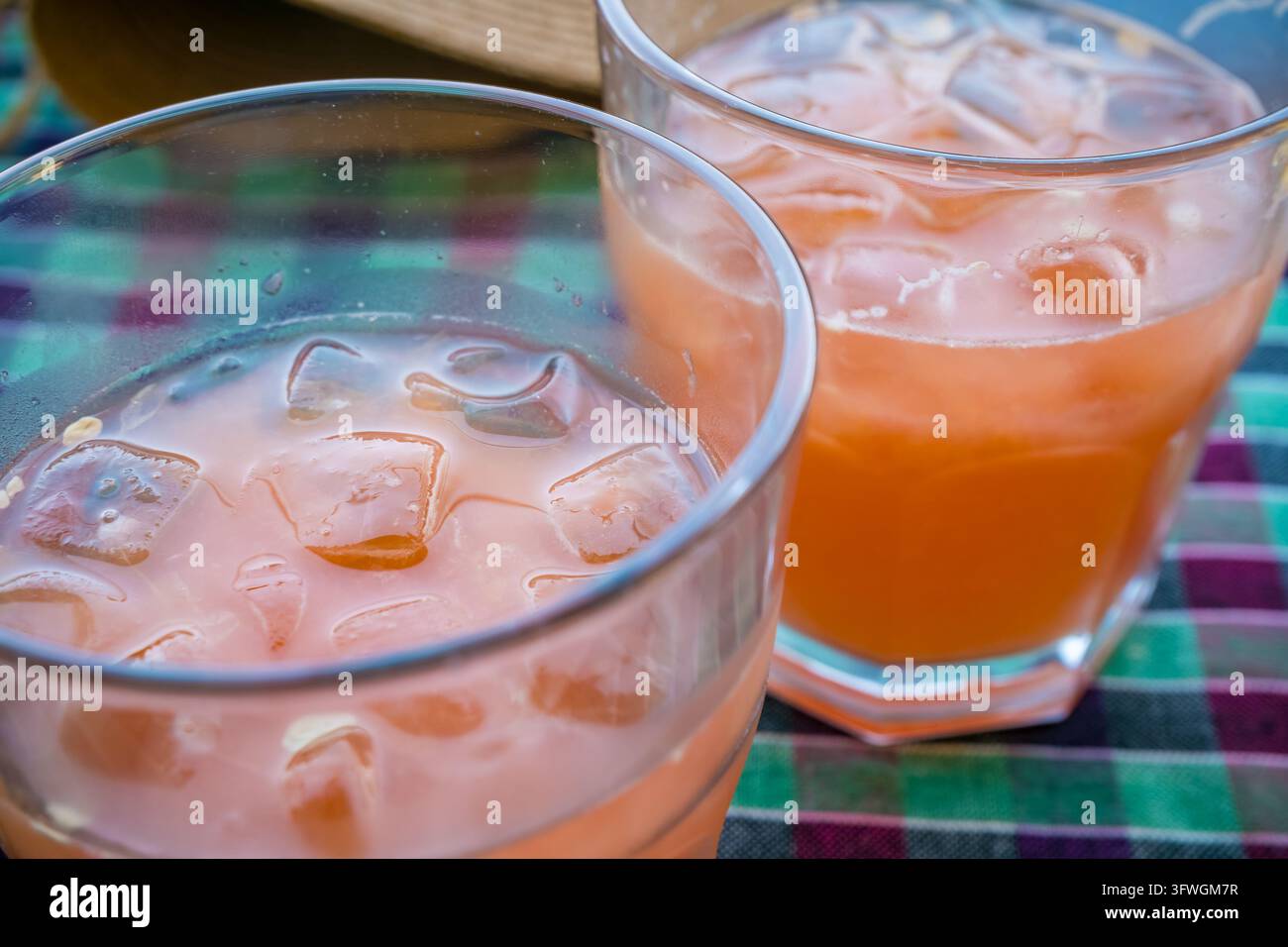 Nahaufnahme von zwei klaren Gläsern mit rosa und orangefarbener Limonade, schmelzenden Eiswürfeln und Kondenswasser auf einer bunten karierten Tischdecke über einem Holztisch Stockfoto