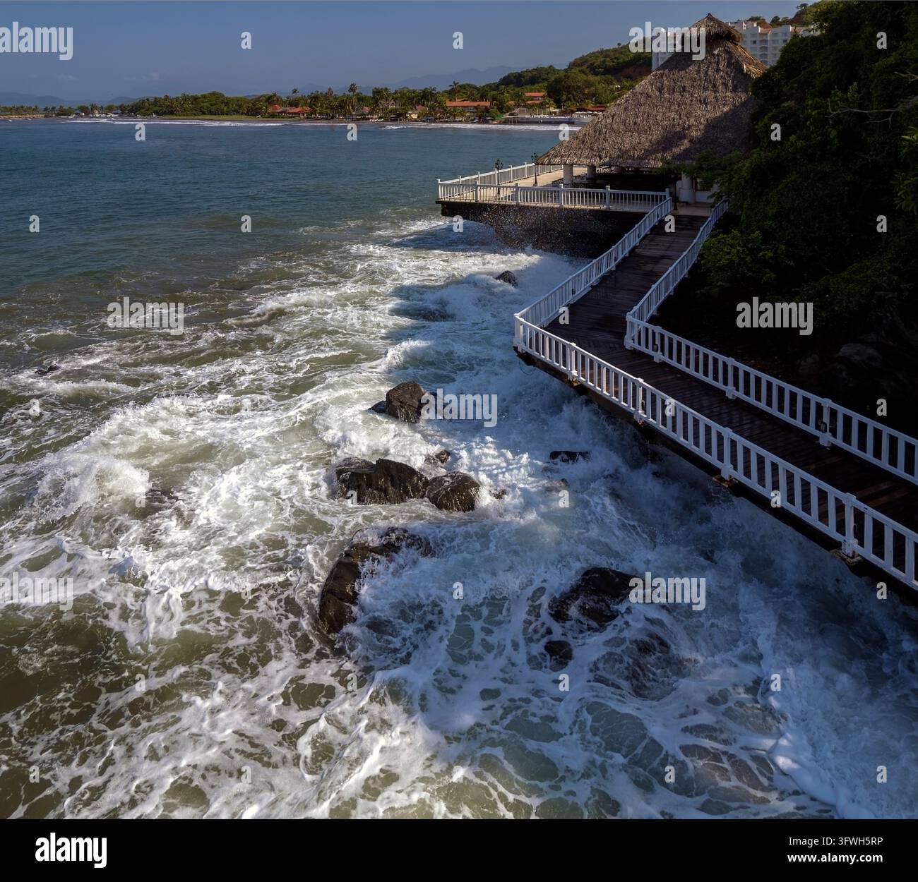 Ein weißer Geländerweg folgt der felsigen Küste neben einer Strohpalapa im Hotel Azul in Ixtapa, Guerrero, Mexiko. Stockfoto