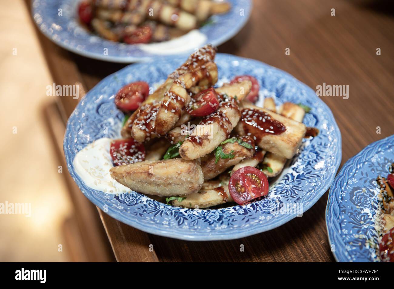 Nahaufnahme eines knusprig gebratenen Auberginensalats, garniert mit Kirschtomaten, süßer Soße und Sesamsamen. Gourmet-Vorspeise auf einem gemusterten blauen Ce Stockfoto