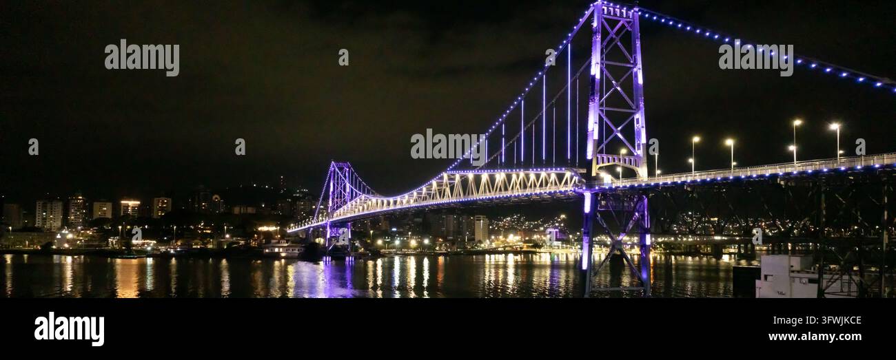 Luftaufnahme der beleuchteten Ponte Hercilio Luz Brücke bei Nacht mit violetten Lichtern, die sich auf dem Wasser spiegeln, und Stadtlichtern im Hintergrund in Flor Stockfoto