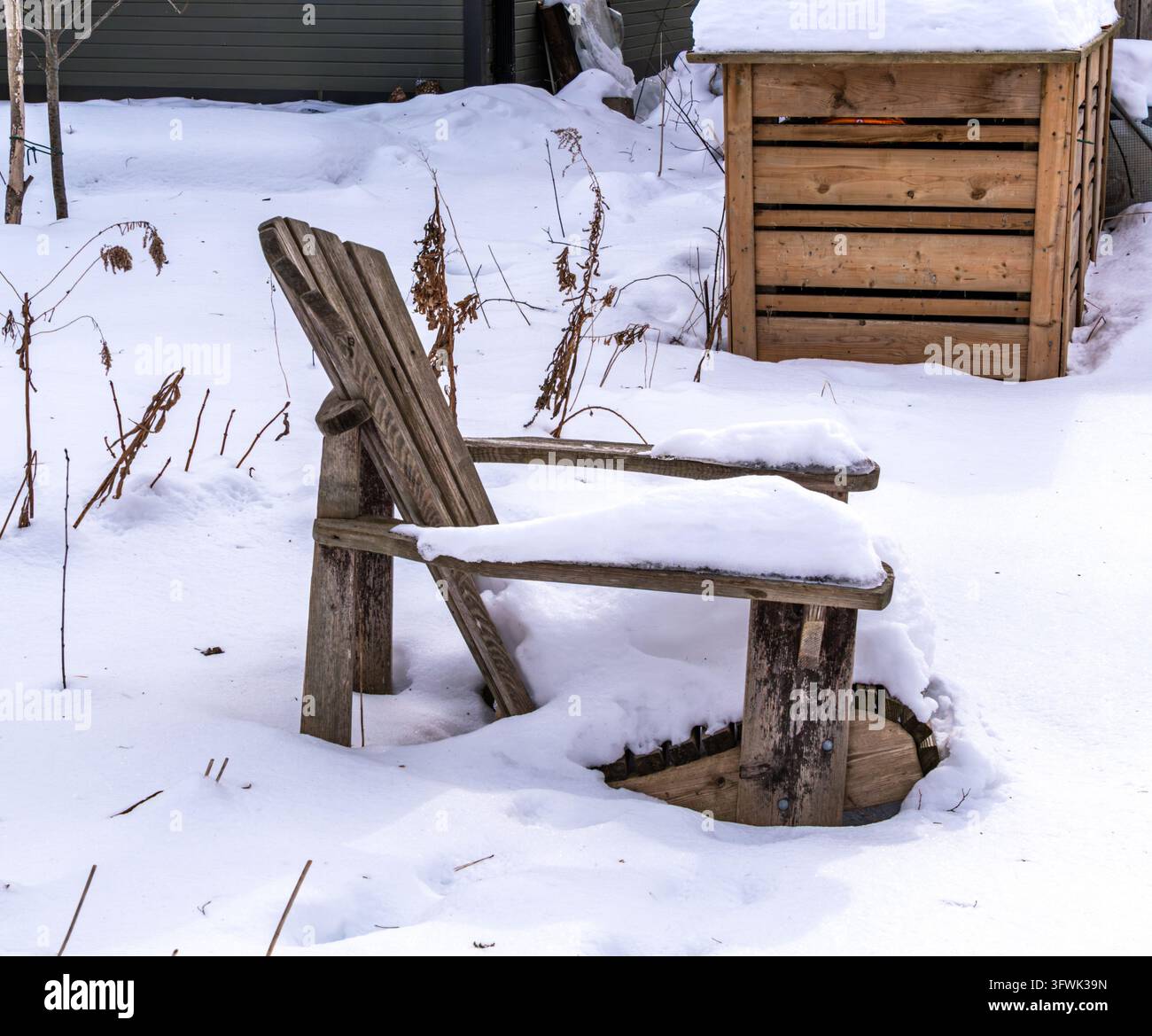 Abgenutzter Holzstuhl, der mit frischem Schnee bedeckt ist, in einem ruhigen Wintergarten, mit getrockneten Pflanzen und einem rustikalen Kompostbehälter im Hintergrund. Stockfoto