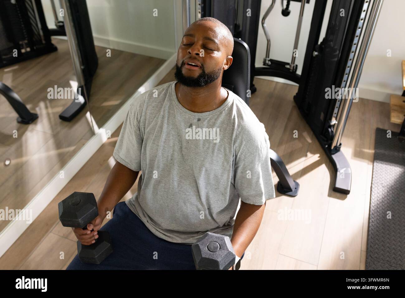 Reifer afroamerikanischer Mann trägt graue T-Shirt-Shorts, sitzt auf einer Bank im Fitnessstudio und hält Sechskant-Hanteln. Stärke, Schweiß, Konzentration, Erholung, Gewichte, Studio, Fitnes Stockfoto